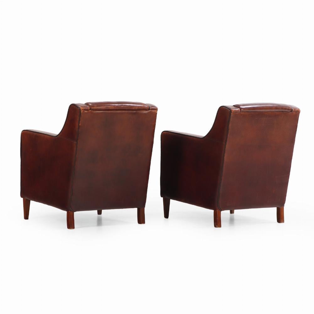 A classic pair of leather club chairs, crafted in Holland circa 1960, featuring warm, aged leather upholstery accented by elegant black piping. The generous proportions, gently squared arms, and supportive backrests offer both comfort and timeless