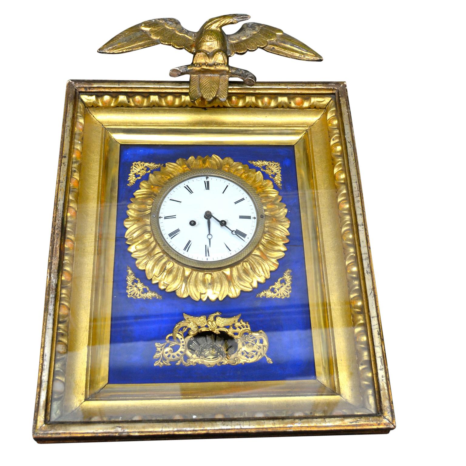 A traditional Austrian Biedermeier wall clock featuring a rectangular carved and gilded wood frame clock face; surmounted by a gilt bronze winged eagle. The decorations on the frame are of gilded metal with gilded carved wood around the dial. The