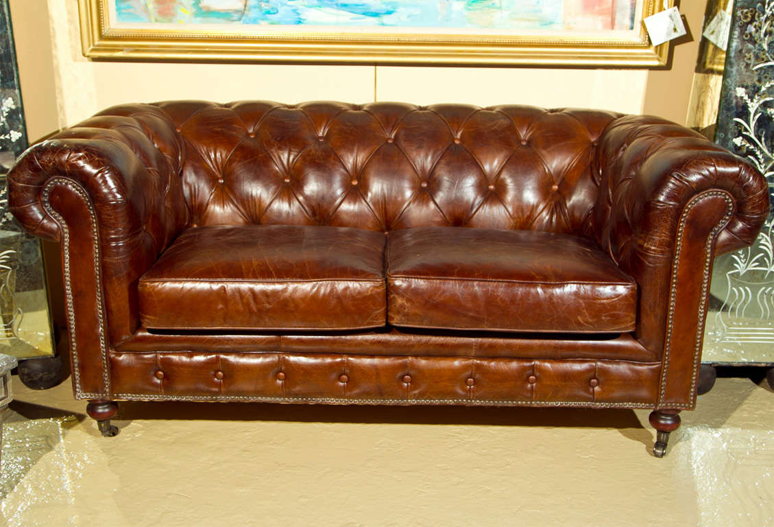 Custom quality English Chesterfield sofa / settee upholstered in worn brown leather, tufted back, rolled arms and cushioned seat, raised on bulbous legs and brass rolling casters. Price is for one. This listing is for a single Chesterfield.