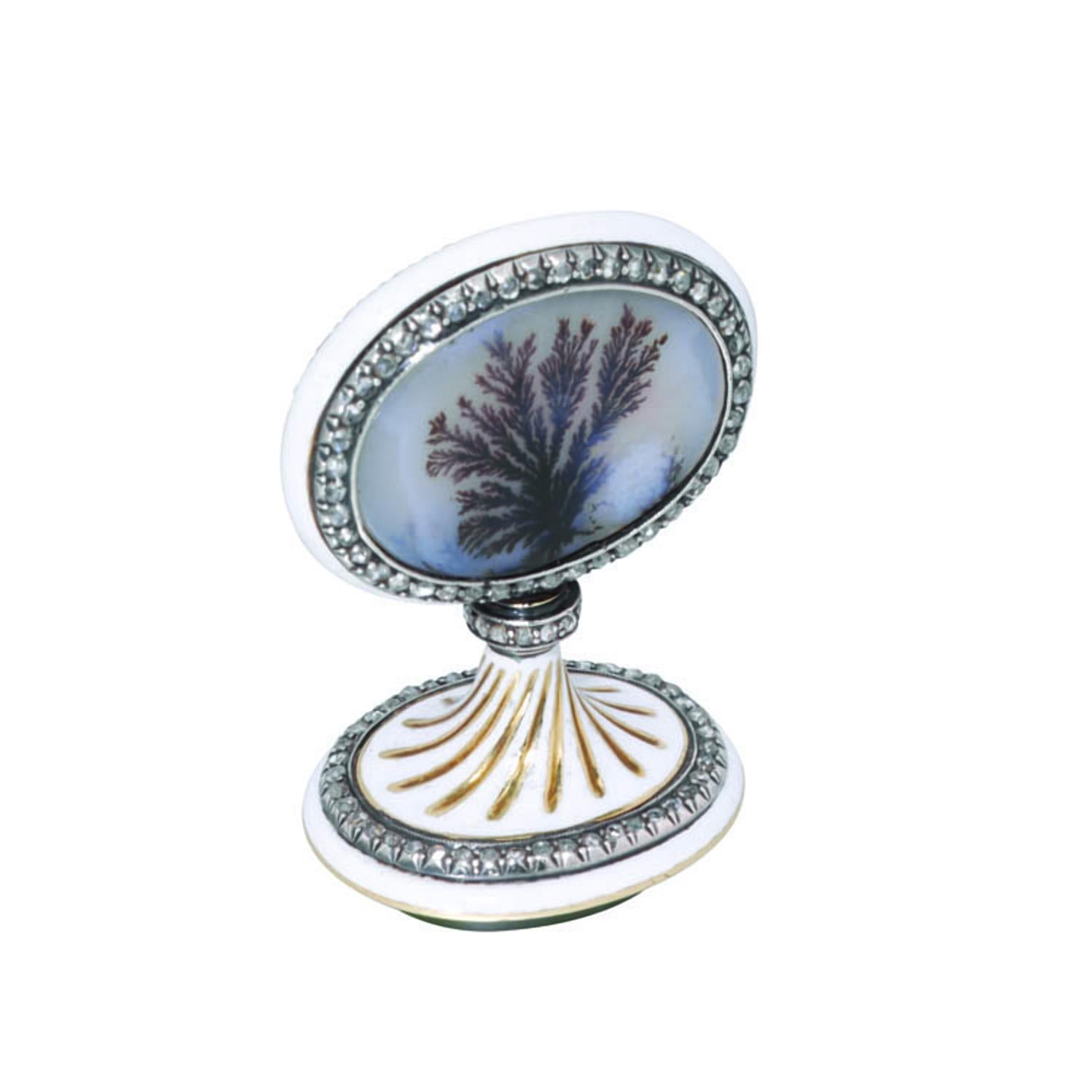 A Faberge' jewelled and enamelled gold and hardstone seal, the flat oval handle inset at both sides with a moss agate panel over sunburst engine-turning within a rose-cut diamond-set border, the mount enamelled in opaque white, the collar with