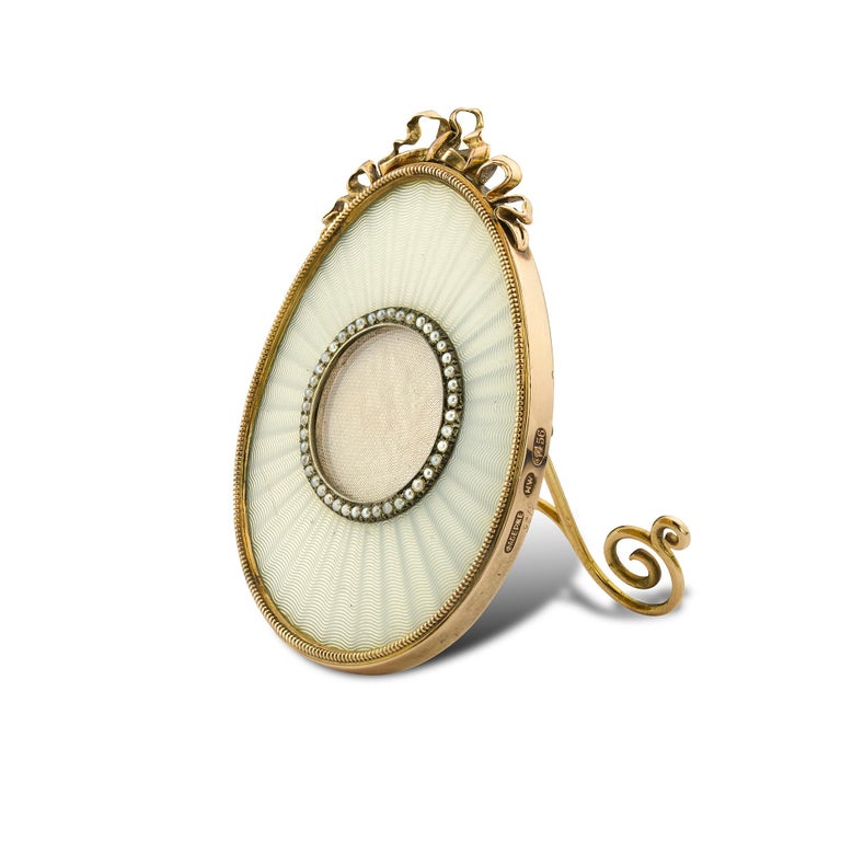 A Fabergé white enamel egg shaped miniature frame, the translucent enamel on an engine turned radiating guilloché ground, with a picture aperture surrounded by 40 seed pearls in a silver setting, the yellow gold outer edge with a ribbon and bow
