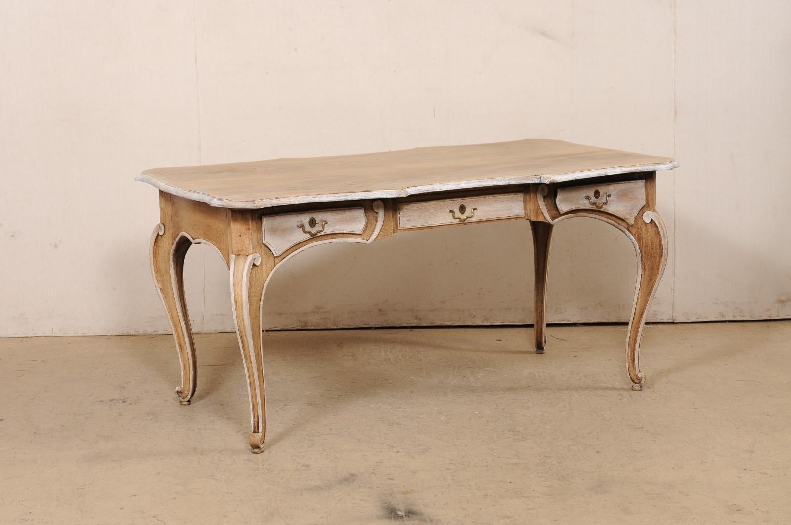 A French bleached-wood desk with three drawers. This vintage table from France features an overall rectangular shaped top which has been nicely emphasized with scallop-carved edges about all four sides, giving this piece a shapely and fluid design