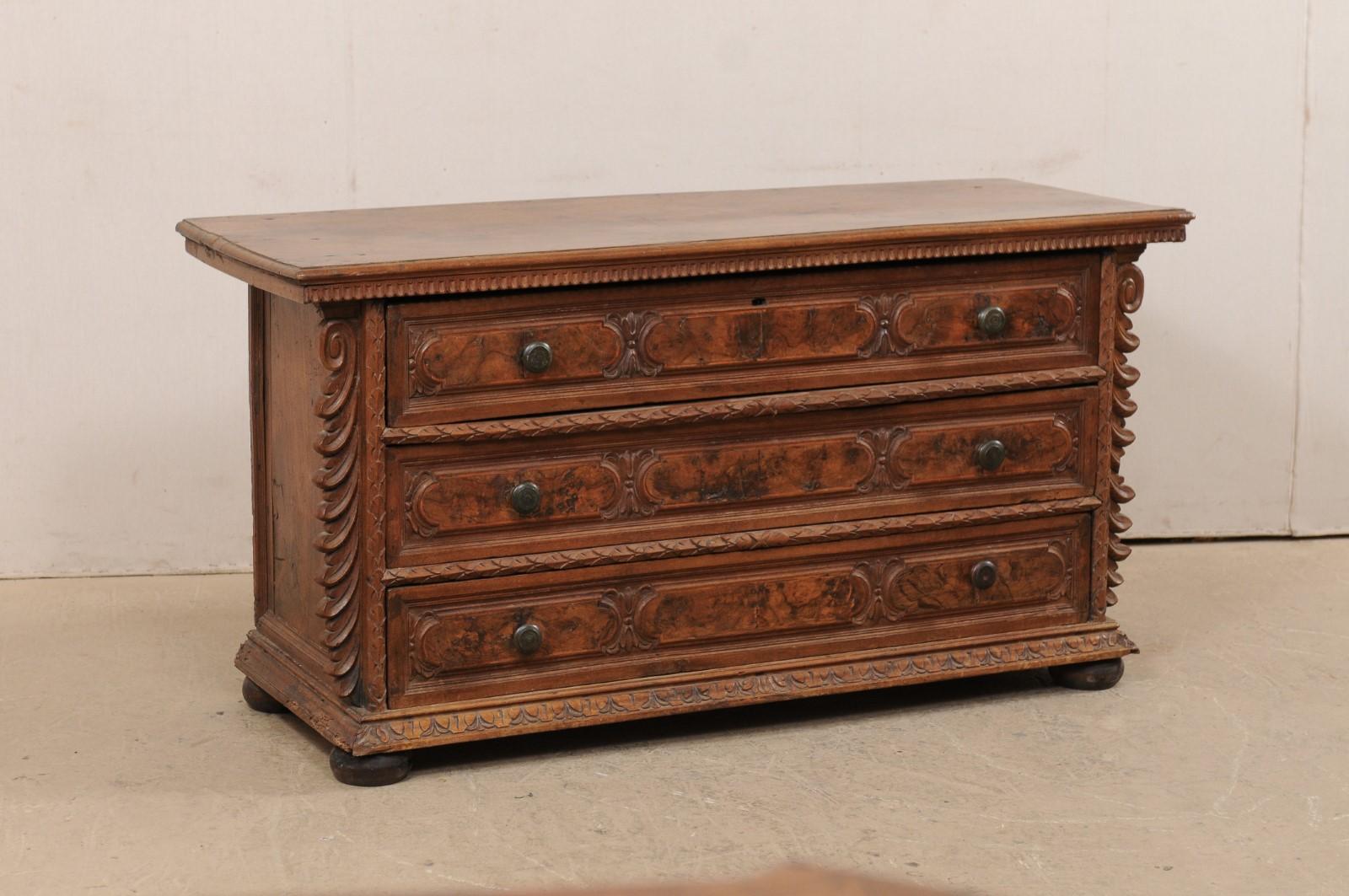 A fabulous Italian carved walnut wood chest of three drawers from the early 18th century. This antique commode from Italy is exquisitely flanked with a pair of carved side posts with volute tops and curled leafy foliage which protrudes outward from