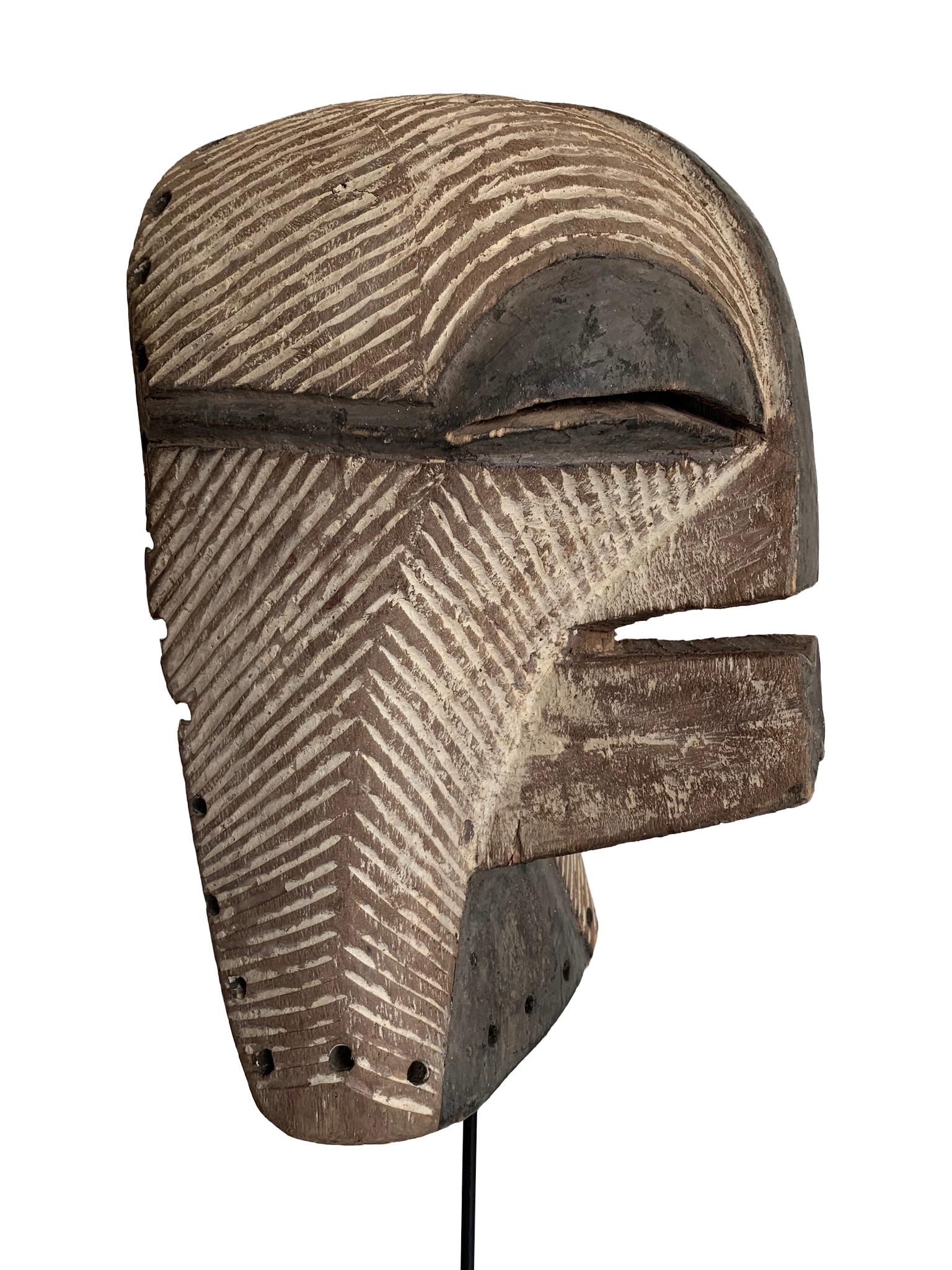 A female Songye Kifwebe hand carved wooden ceremonial mask. The wooden mask is carved with geometric lines on the face and head which are whitened with pigment and darkened on the large ridged nose, mouth and eyes. Around the back edge are the holes