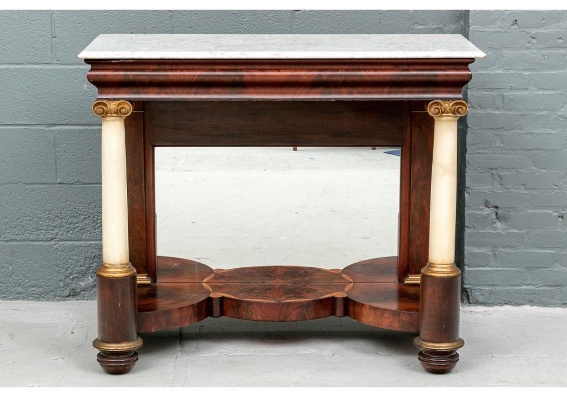 A 19th century classical Empire pier console table having a beveled rectangular marble top above an Ogee mahogany veneered apron raised on carved and gilt white marble Ionic column supports. The unusual shaped serpentine plateau before an inset