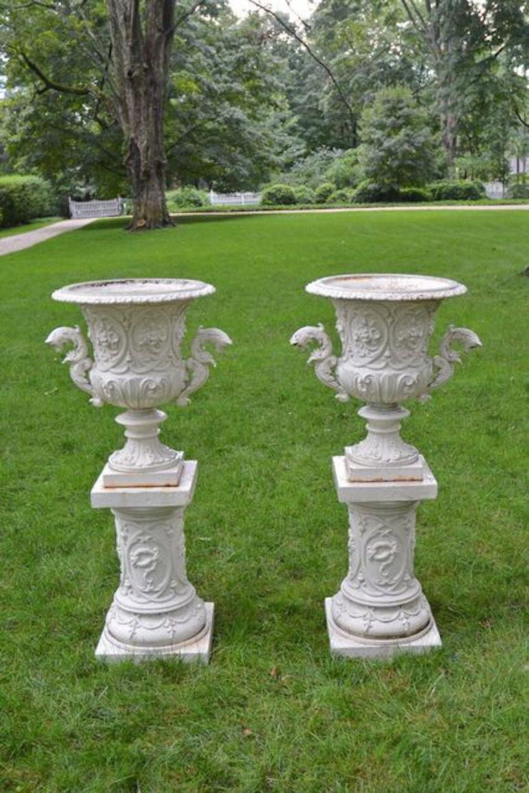 An elaborate rare pair of cast-iron urns on pedestals, each campana form urn in the Rococo Revival style, with interlocked scrolls and floral cornucopia motifs, with scrolled and foliated animal-form handles. Raised on cylindrical pedestals, the