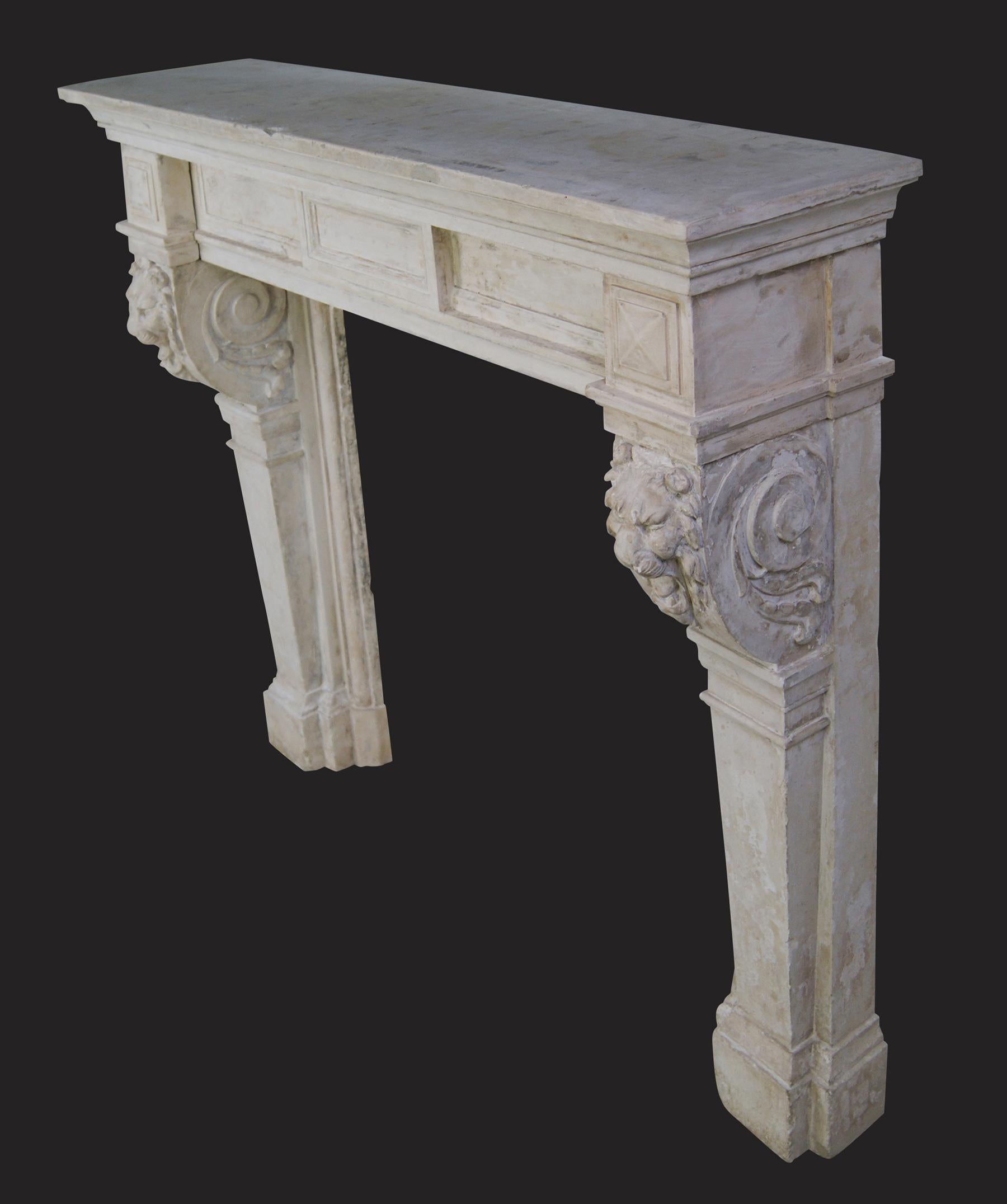 Fina chimenea antigua de piedra, de forma arquitectónica, con jambas de columnas cónicas platinadas que sostienen ménsulas de roleos con frente de león finamente talladas.  Los esquineros romboidales flanquean el sencillo friso con tablilla central