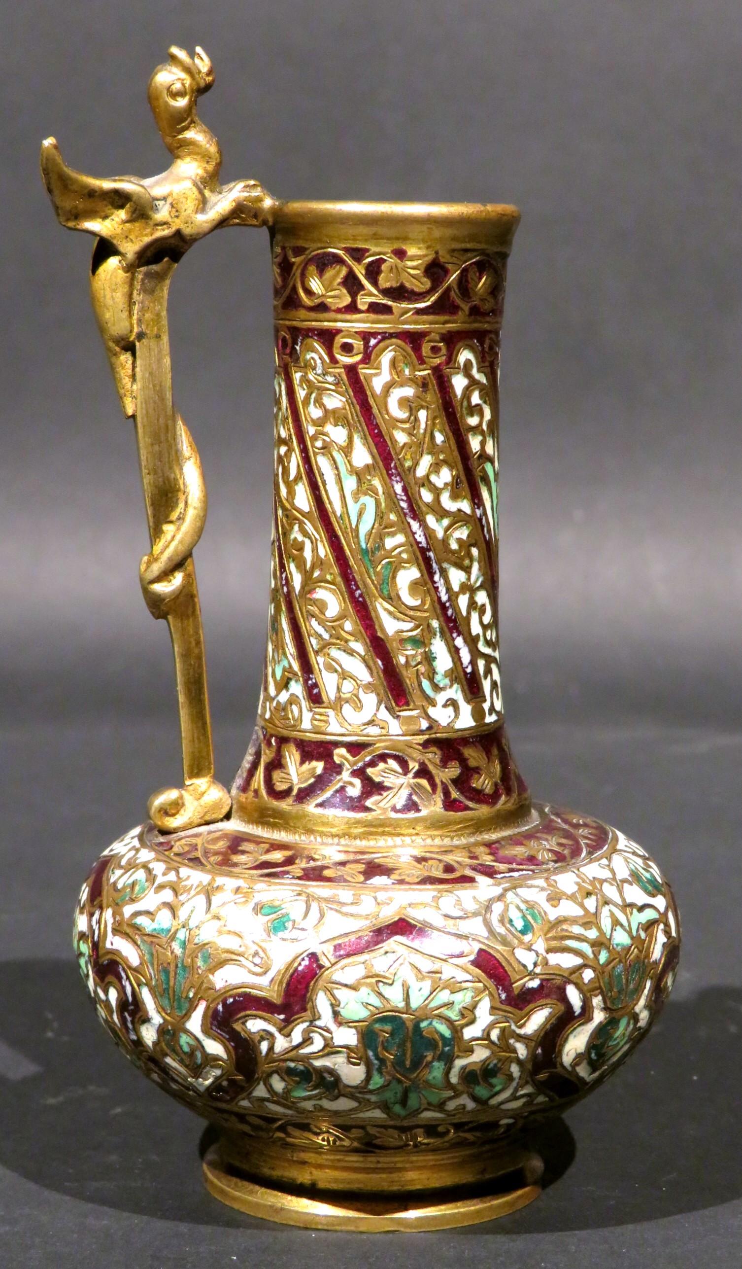 The stunning & richly gilded bronze body profusely decorated overall with hand applied, jewel-like arabesque motifs in a palette of multi-coloured enamels, the flaring columnar neck further decorated with an applied gilt bronze handle in the form of