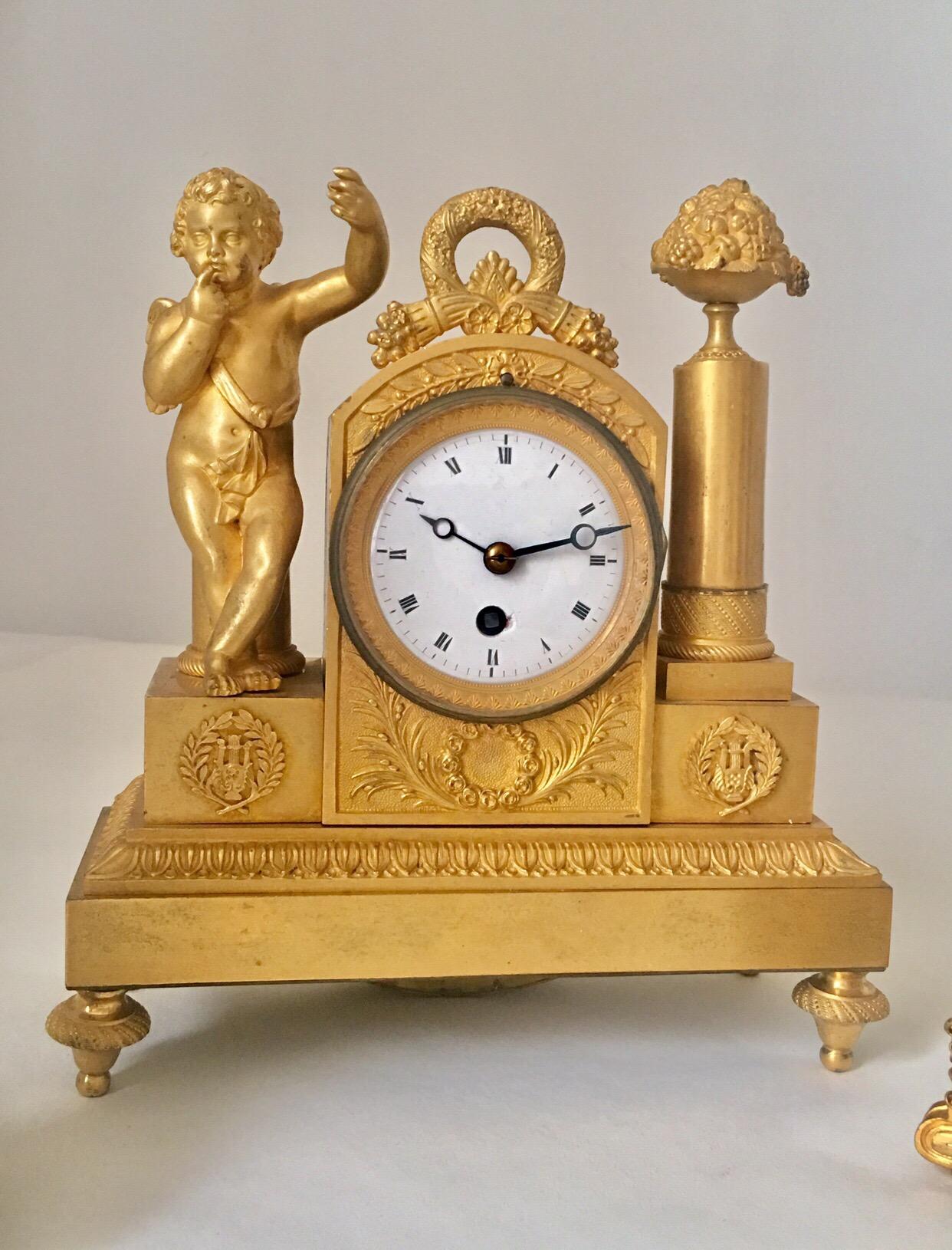 A very fine early 19th century miniature French Empire period 3-pieces set cherub clock

The clock with an arched case raised on a plinth base and four toupie feet, mounted with a figure of cupid to one side and a cylindrical pedestal supporting a
