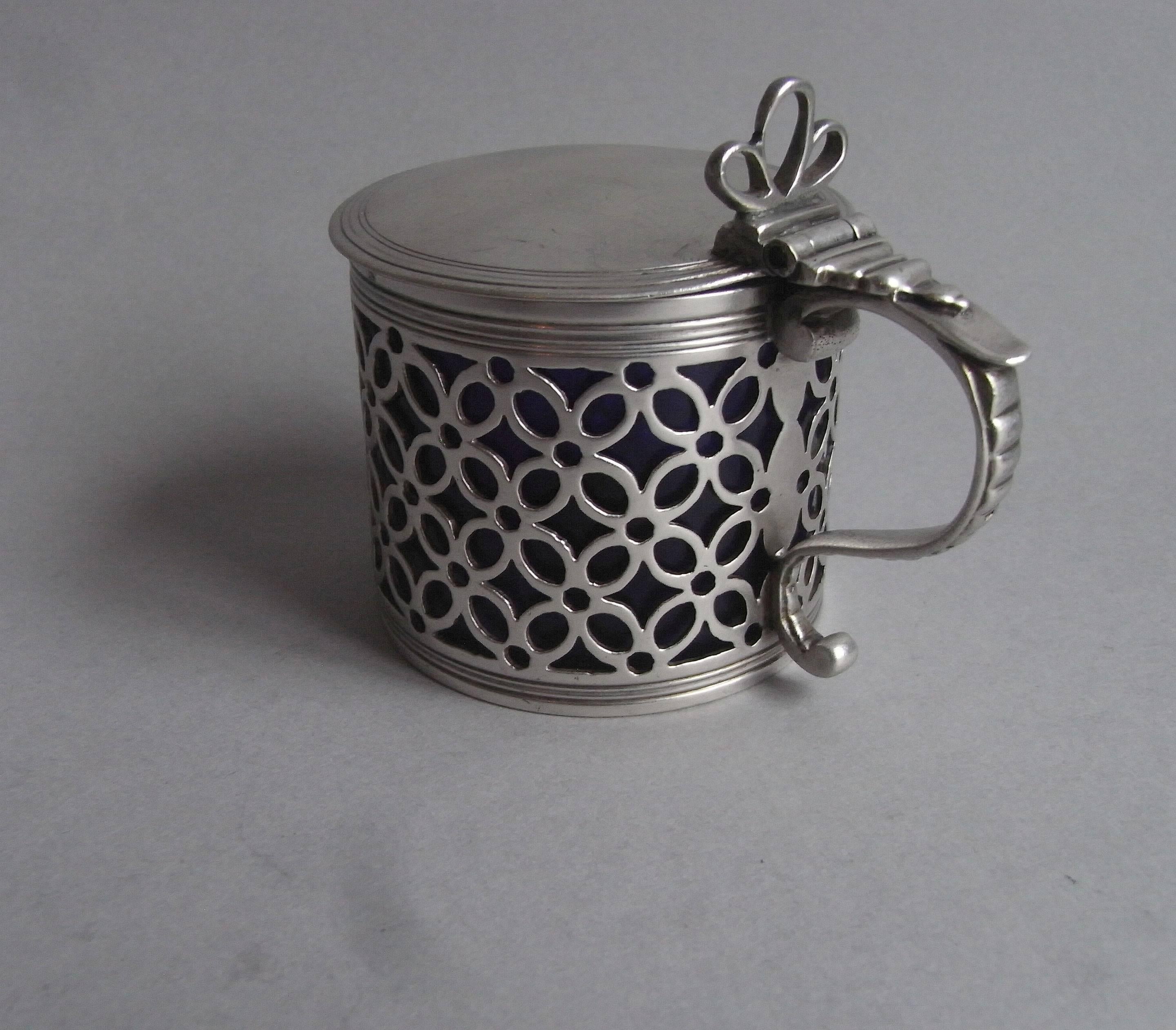 The mustard pot is modelled in the drum form and the sides are pierced with quatrefoil designs and roundels. The cover displays a reeded edge and the scroll handle is decorated with ribbing. This piece has a tri-furcated pierced thumb piece and a