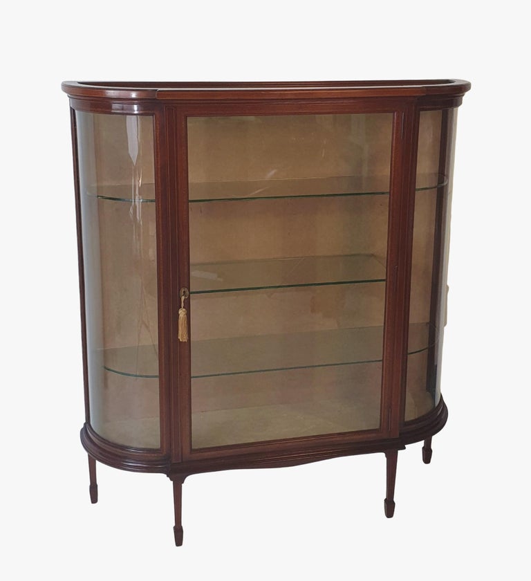 Early twentieth century inlaid mahogany display cabinet. The moulded bow front top raised above centred glass door with brass escutcheon and key flanked by two shaped glass panels enclosing a three shelved interior supported on shaped apron above