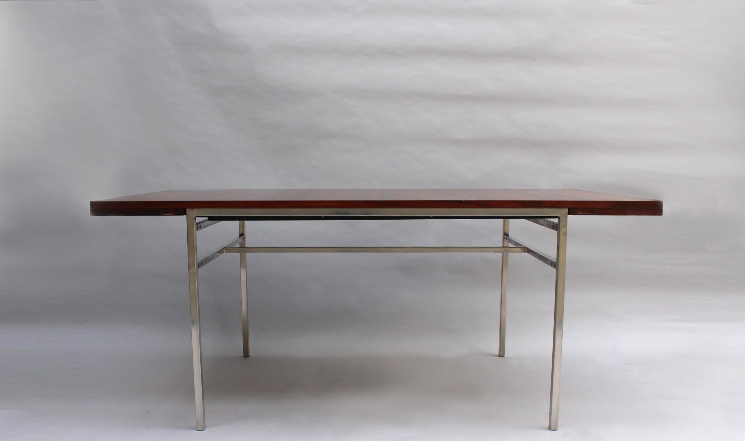 A fine French midcentury rectangular dining table with a chromed metal base, veneered rosewood top and 2 foldout matching end leaves (length with extensions is 94.5 inches). Labelled.