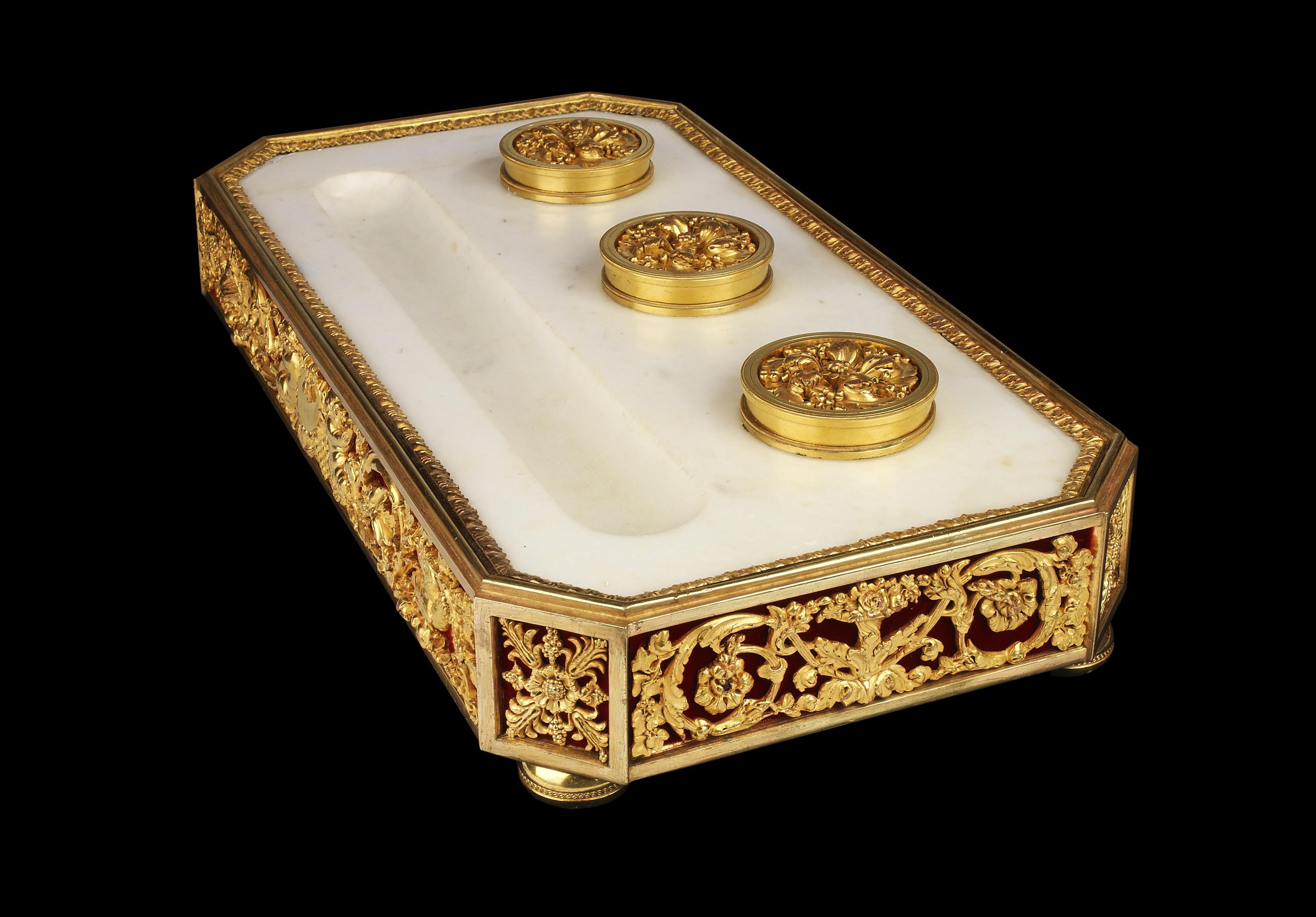 Rectangular white statuary marble inkstand with elaborate gilt bronze mounts applied to the sides backed by red velvet. The surface has three ink containers with ormolu lids and a cutout valley for the pens, resting on ormolu turned bun feet.
