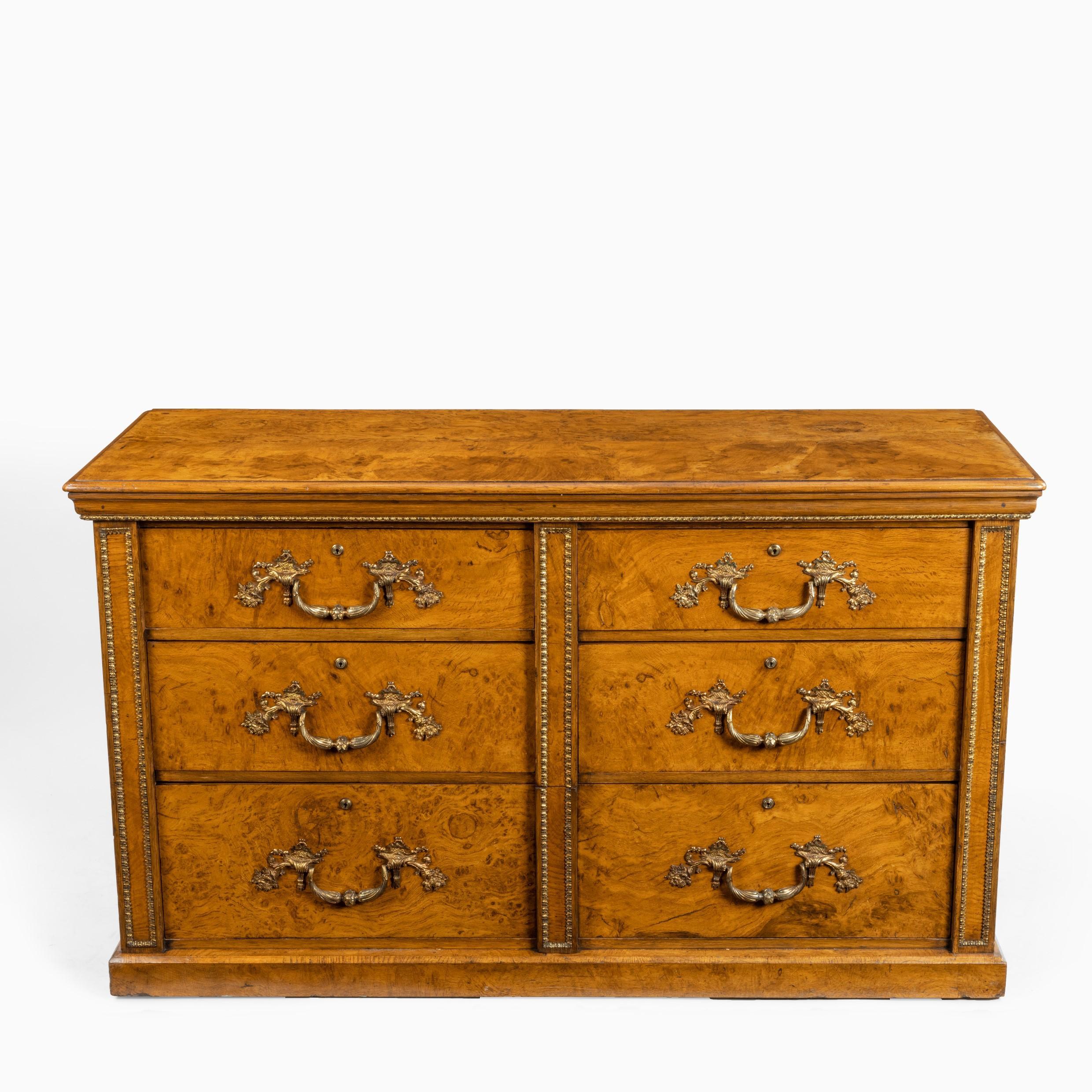 A fine George IV burr oak chest of drawers in the manner of Morel and Seddon, of rectangular form with three tiers comprising four short and one disguised long drawer, on a plinth base, decorated with ormolu edging and the original Rococo handles.