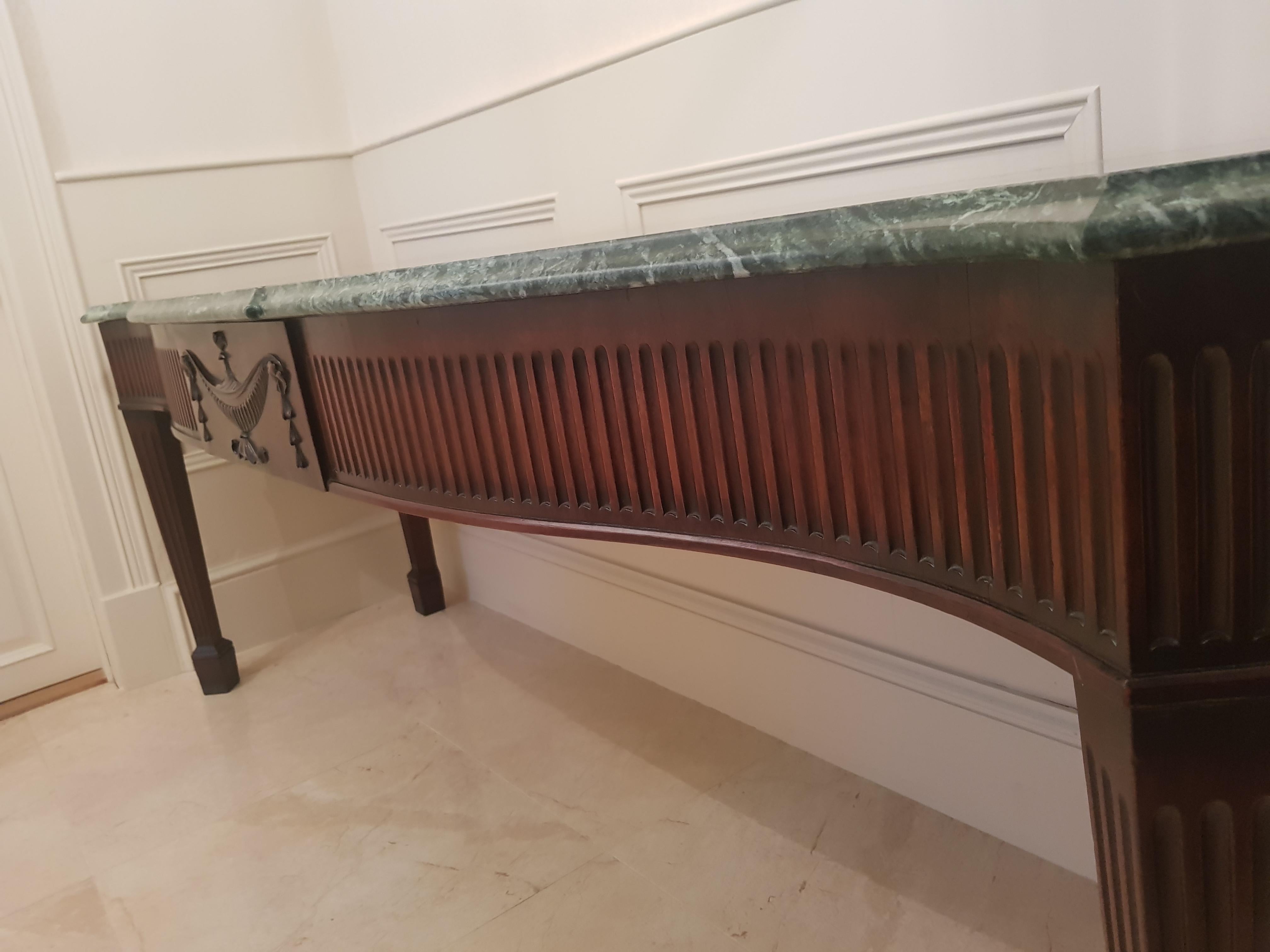 A fine mahogany serving table, with moulded breakfront and serpentine green verde marble top with canted corners, above a deep fluted frieze centred with a swagged urn with a flower finial, on square fluted legs with moulded blocked feet. Irish