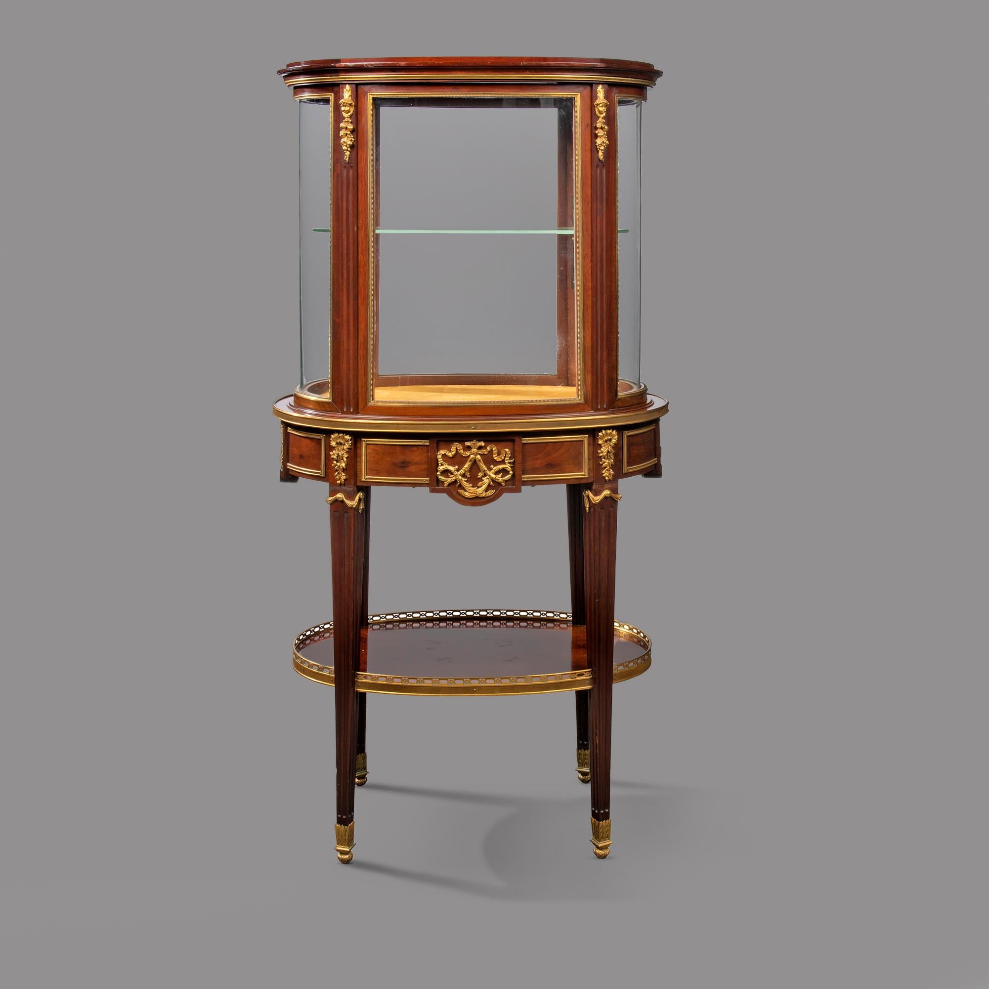 A Fine Louis XVI Style Gilt-Bronze Mounted Mahogany Centre Display Cabinet or ‘Vitrine de Milieu’, By Paul Sormani, Paris. 

The oval vitrine cabinet with four curved glass panes divided by fluted pilasters with cherub mask fruiting gilt-bronze