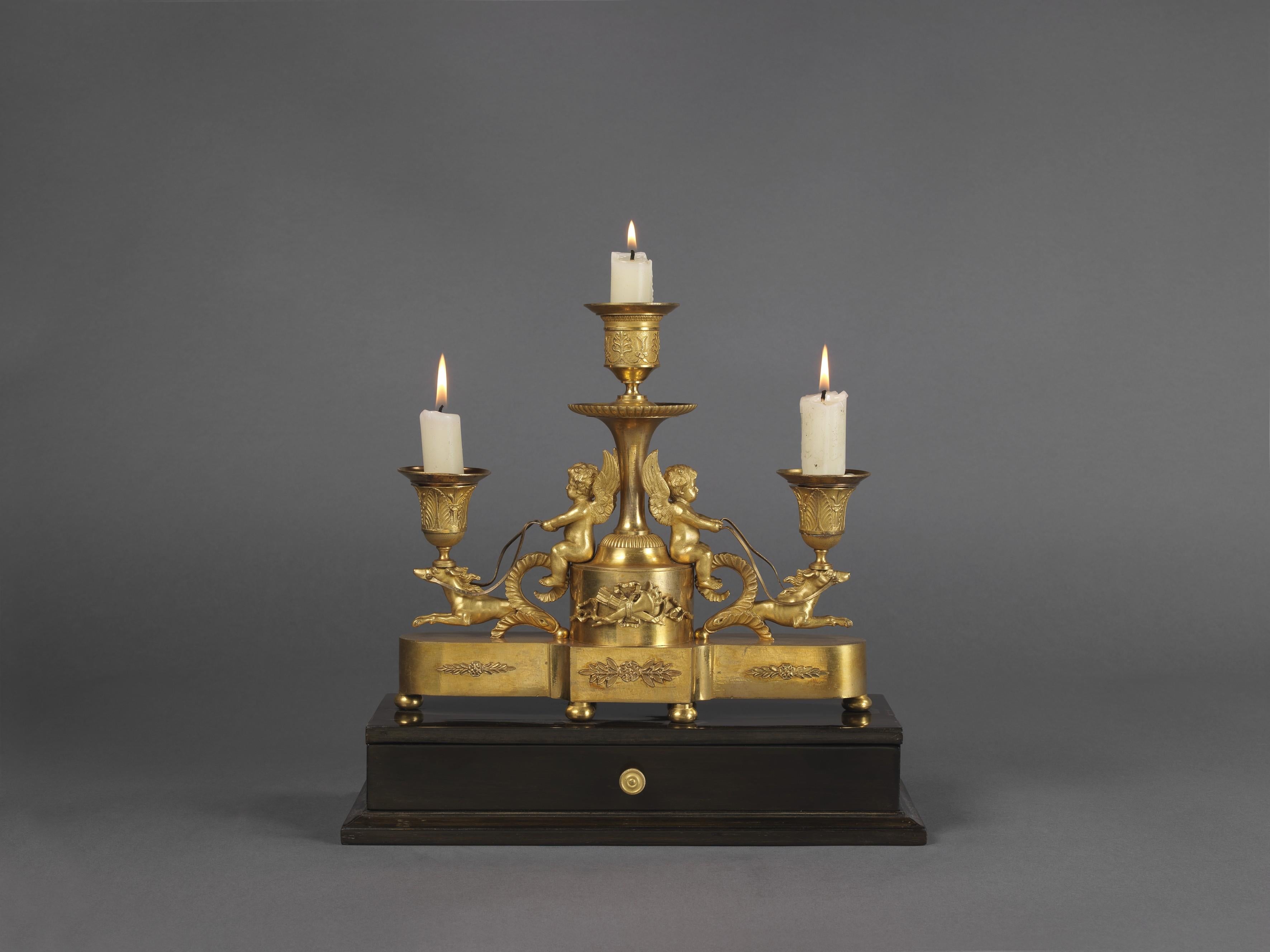 A fine Louis XVI style gilt bronze and ebony three-light desk stand.

French, circa 1820. 

This elegant desk stand has an ebonized rectangular base with drawer, supporting a re-entrant gilt-bronze plinth with ball feet. The plinth supports a