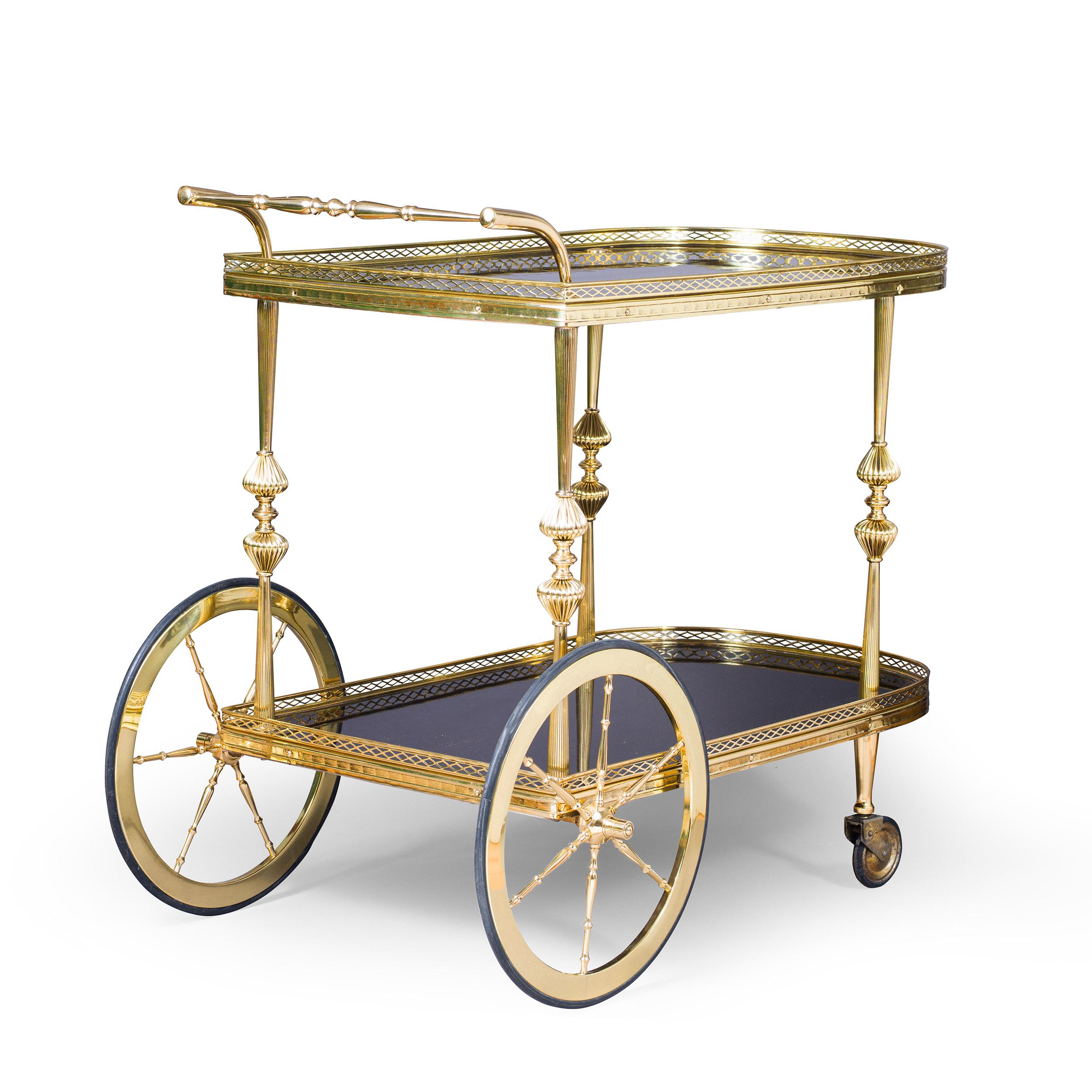 A fine mid-century drinks trolley or bar cart, the two tiers with black polished tops within polished brass pierced galleries, the top supported on fluted brass legs with cast details. With large polished wheels below the handle and smaller trolley