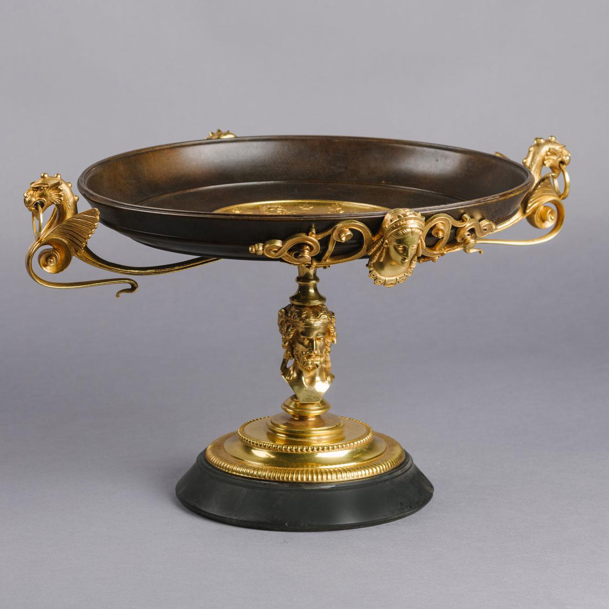 A fine neoclassical Revival gilt and patinated bronze Tazza.

The circular dish centred by a gilt-bronze roundel of the Greek God Zeus enthroned, flanked by chimerical handles and raised on a figural stem with a spreading stepped foot, on a grey