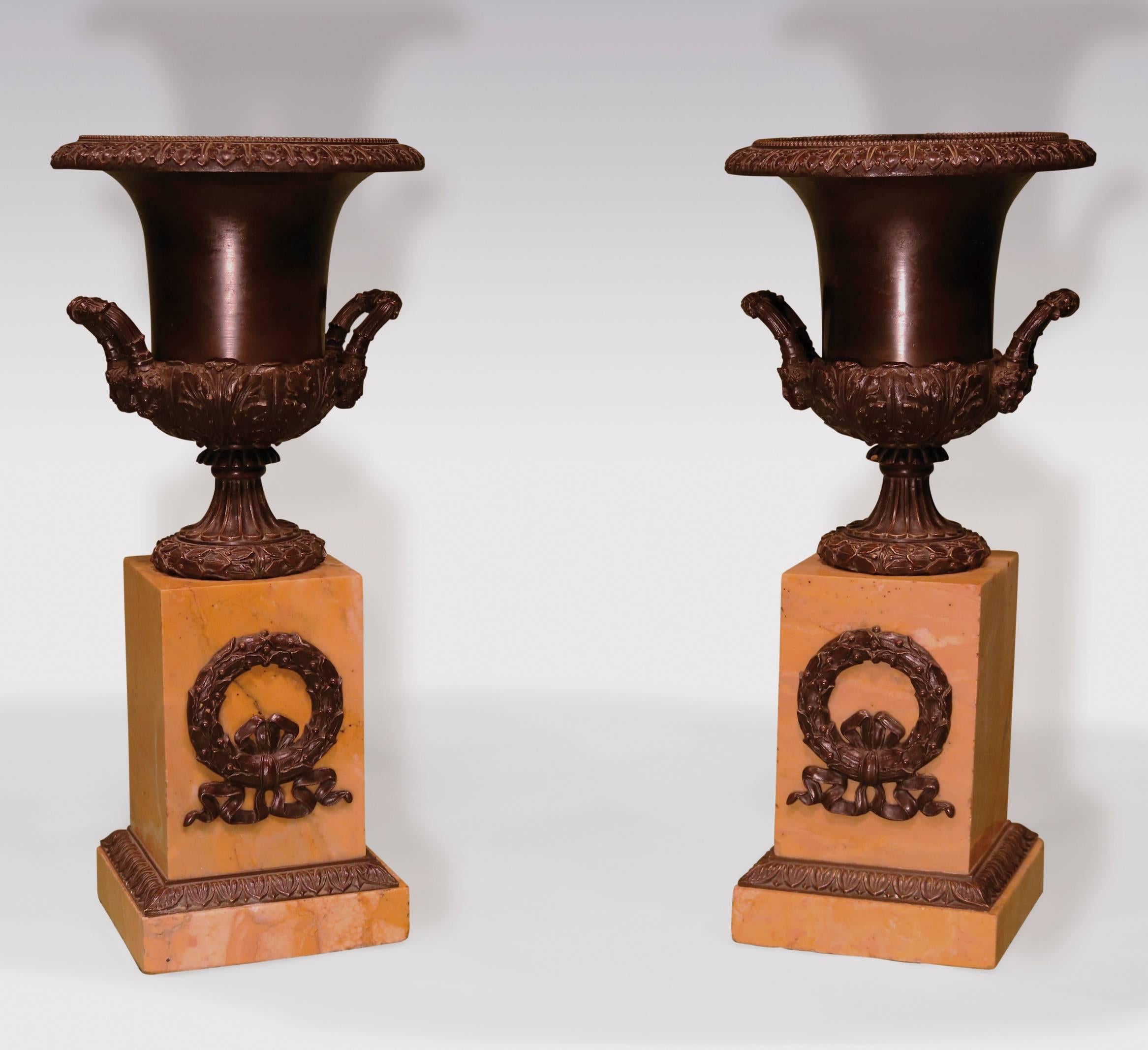 A pair of early 19th century bronze campana-shaped urns, having leaf and berry beaded rims, above scrolled satyr mask handles, raised on fluted stems, supported on sienna marble plinth bases, with ribbon-tied wreath appliqués. (Now converted to