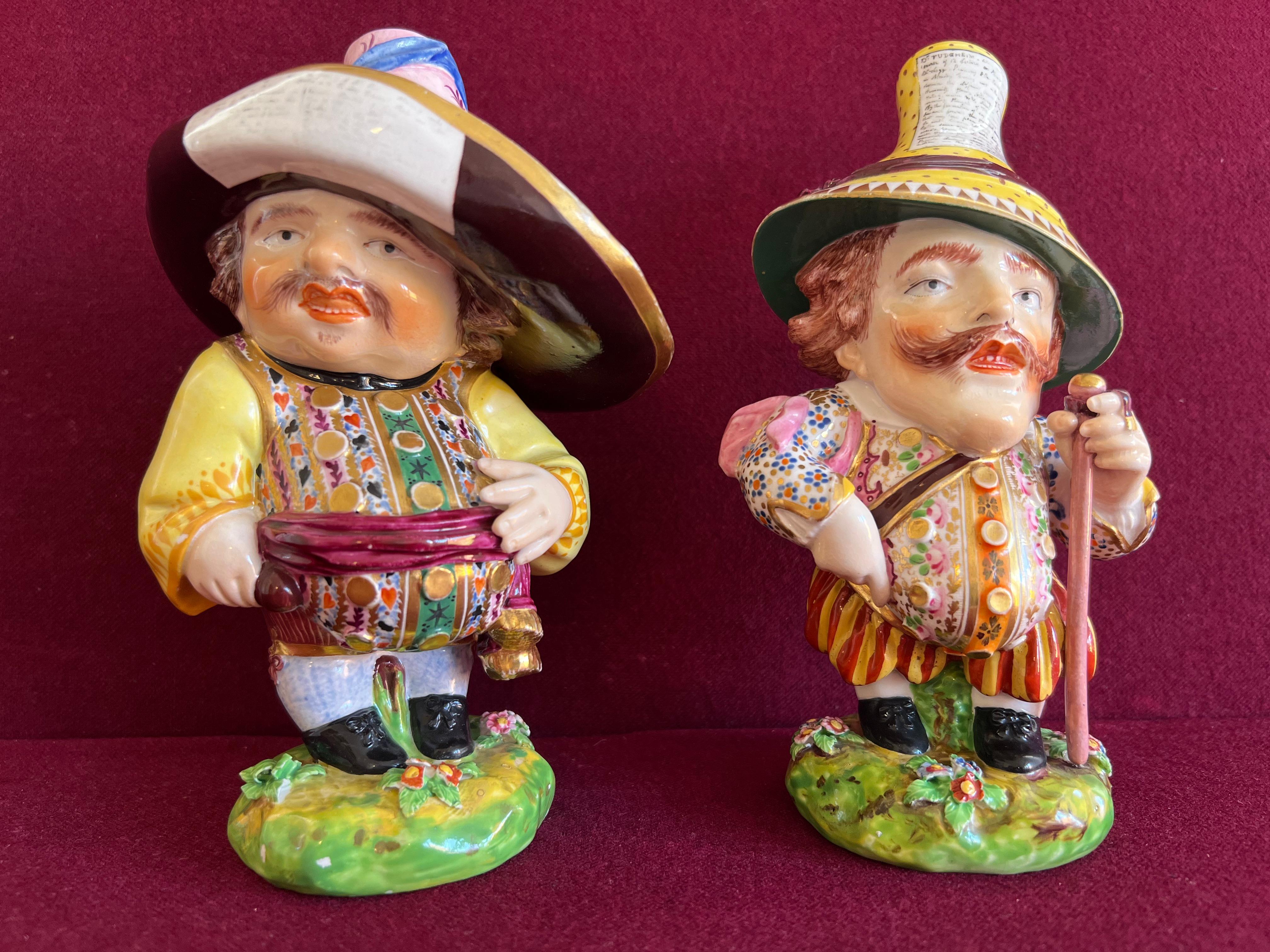 A fine pair of Bloor Derby ‘Mansion House’ Dwarfs c.1825. Each moustached figure modelled wearing a comically large hat bearing an advertisement, one wearing a tasselled sash about his rotund belly, the other with polka dots on his shirt sleeves, a