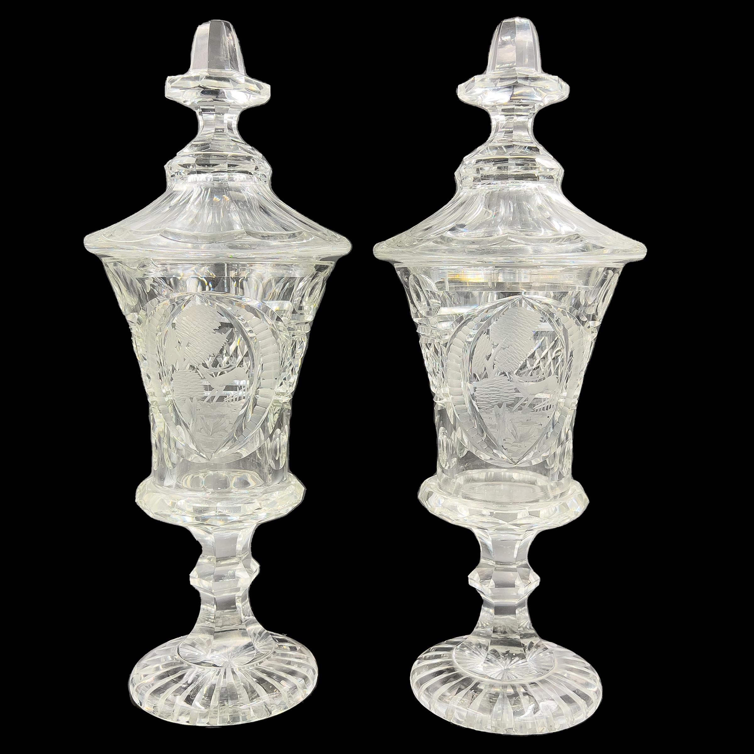 Une paire de vases en verre clair de Bohème, chacun avec un couvercle bombé évasé surmonté d'un fleuron octogonal, des scènes de faune et de flore à travers un paysage, le tout reposant sur une tige hexagonale à boutons se terminant par un pied