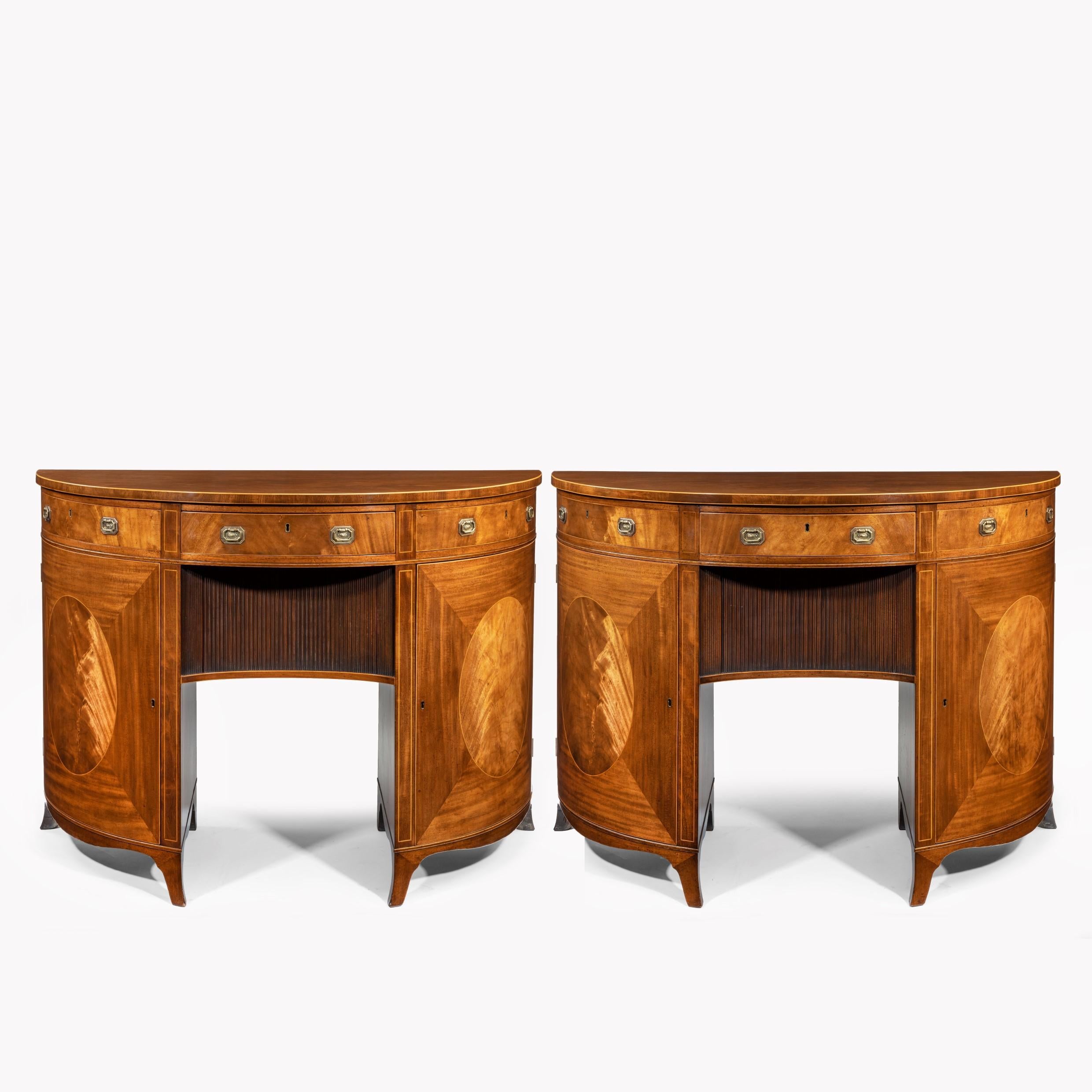 A fine pair of George III figured mahogany side cabinets, in the manner of Thomas Sheraton, each demilune top with a strung edge above a central frieze drawer flanked by dummy drawers, all above a shaped tambour door between shaped cupboard doors