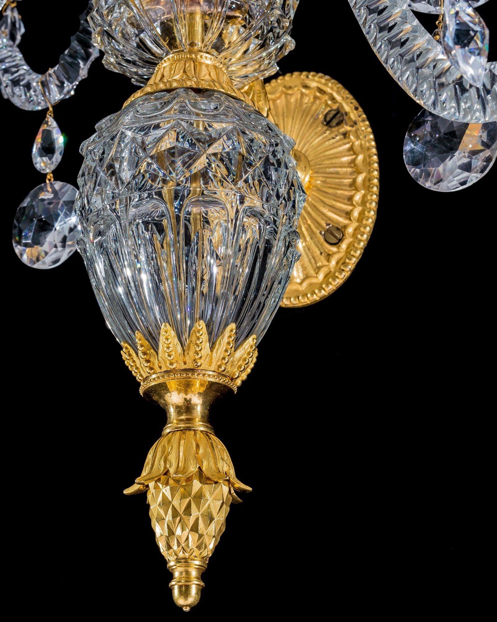 A fine pair of ormolu and cut glass wall lights suspended by oval fluted back plates with ball bead decoration, mounted with a scroll arm supporting cut glass eggs, mounted with a ormolu pineapple and foliate socle. The diamond cut container issuing