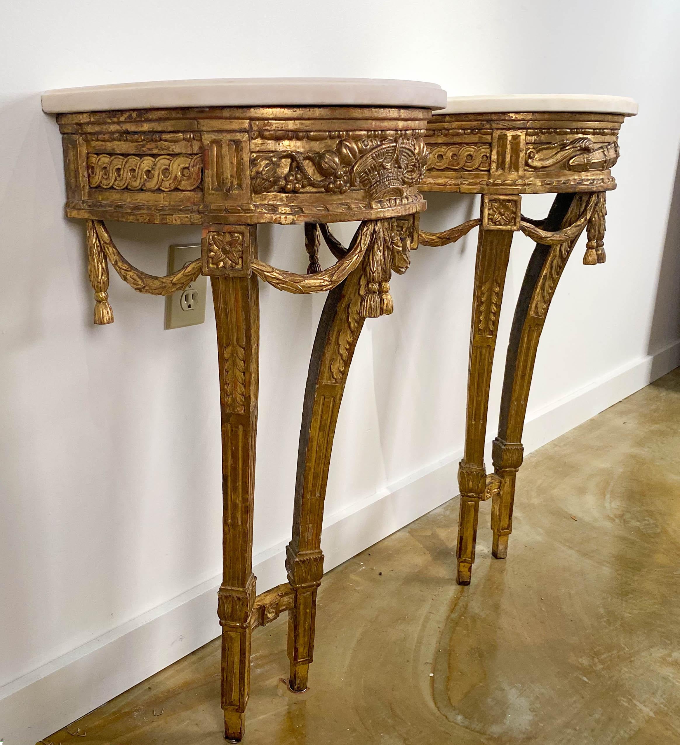 A fine pair of small Louis XVI 
Carved giltwood console tables 
18th century

Each with a D-shape white Carrara marble top,  above the guilloche-carved frieze, one centered by a woven basket flanked by fruit, the other with a sheaf of arrows.  Both