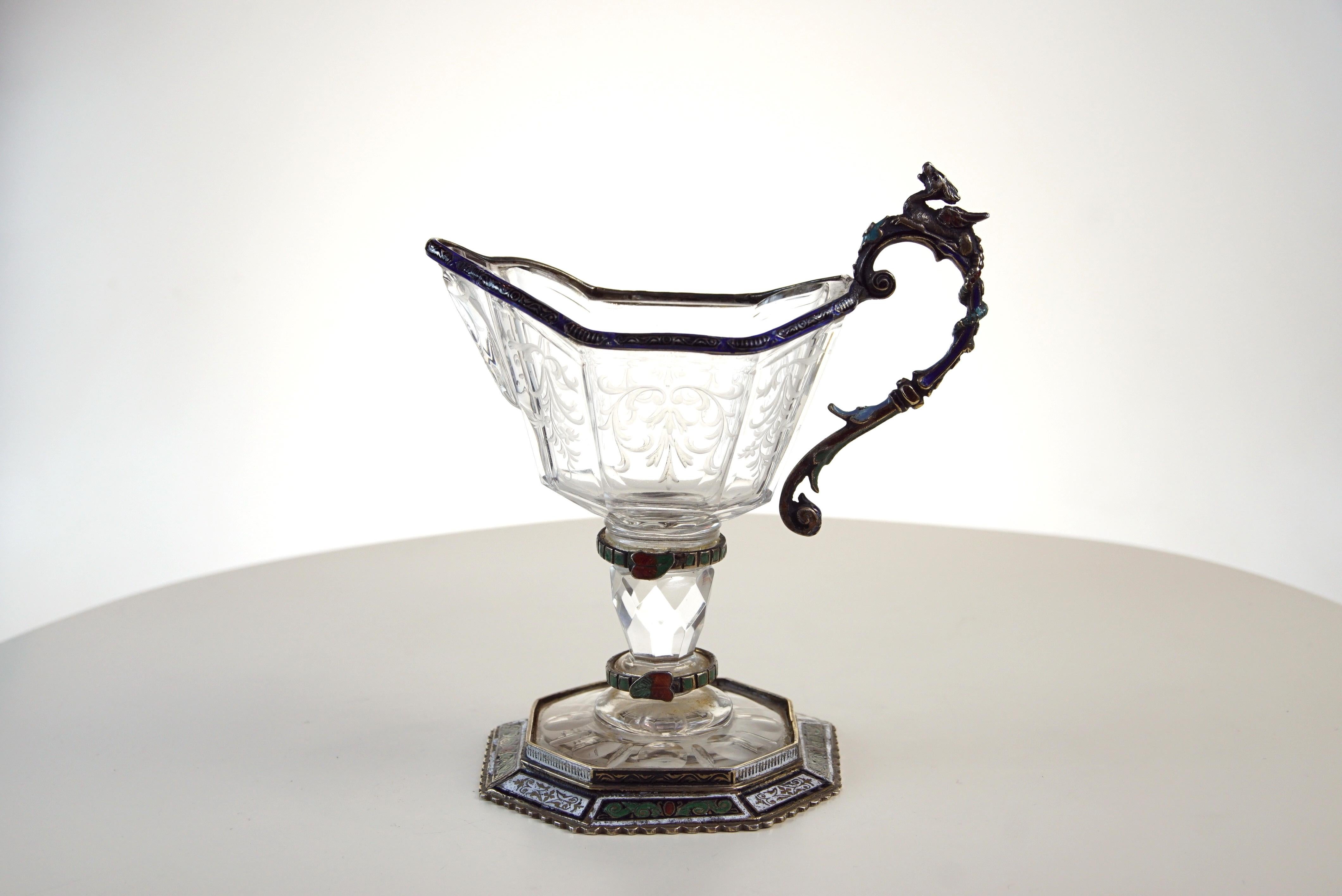 A fine silver, enamel, and engraved rock crystal jug, circa 1880,
attributed to Hermann Bohm

The rock crystal body engraved with scrolls ,The handle with an enameled mystical Bird supported by a rock crystal column on an engraved rock crystal
