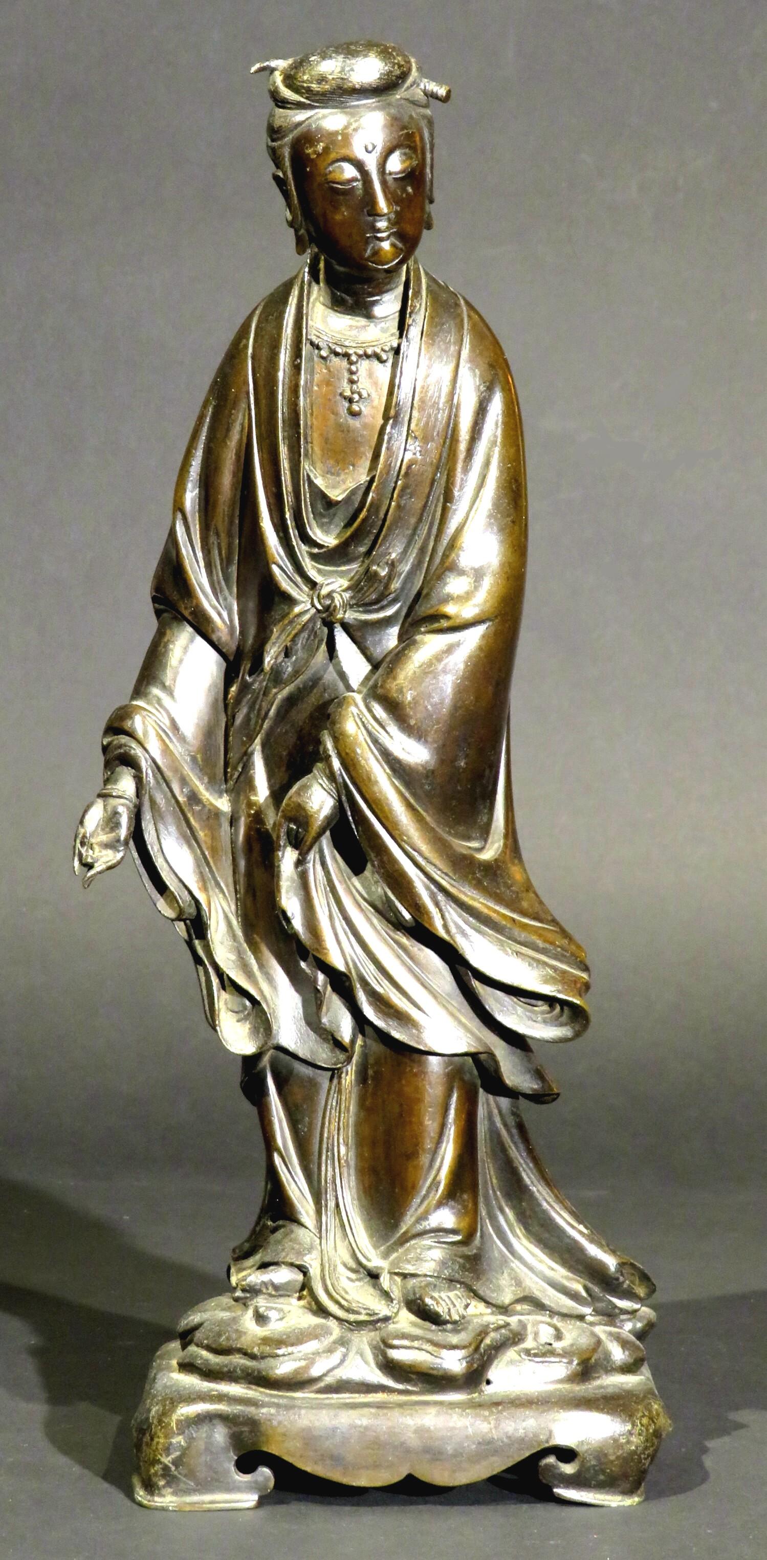 The finely cast bronze figure of Guanyin - Bodhisattva Goddess of Compassion, Kindness & Mercy. 
Shown standing atop a naturalistic mound on a scrolled rectangular base, wearing an outer robe with finely modelled cascading folds tied at her waist,