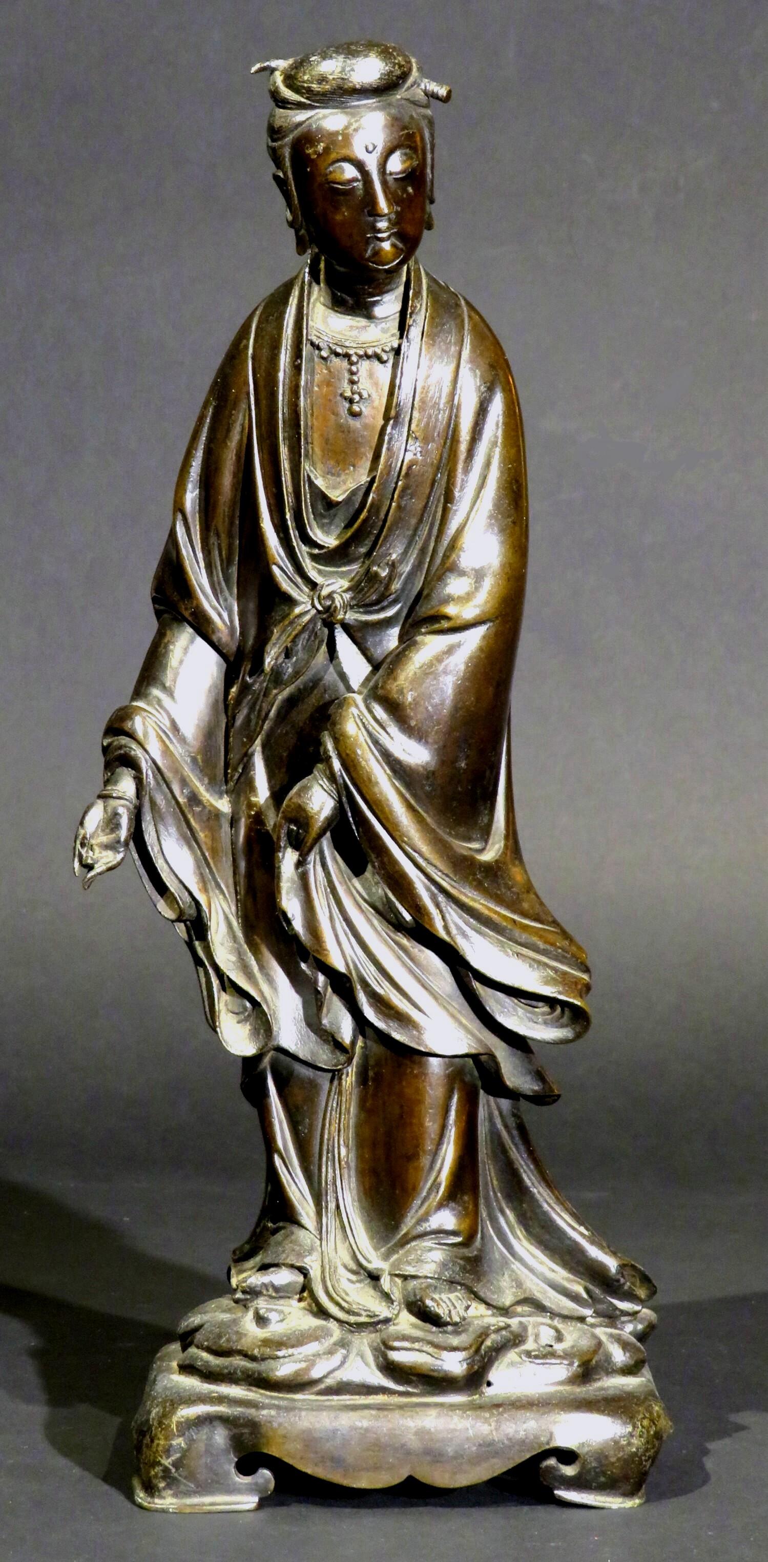 The finely cast bronze figure of Guanyin - Bodhisattva Goddess of Compassion, Kindness & Mercy. 
Shown wearing an outer robe finely modelled with cascading folds tied at her waist, her head subtly tilted to the right, her face with downcast eyes,