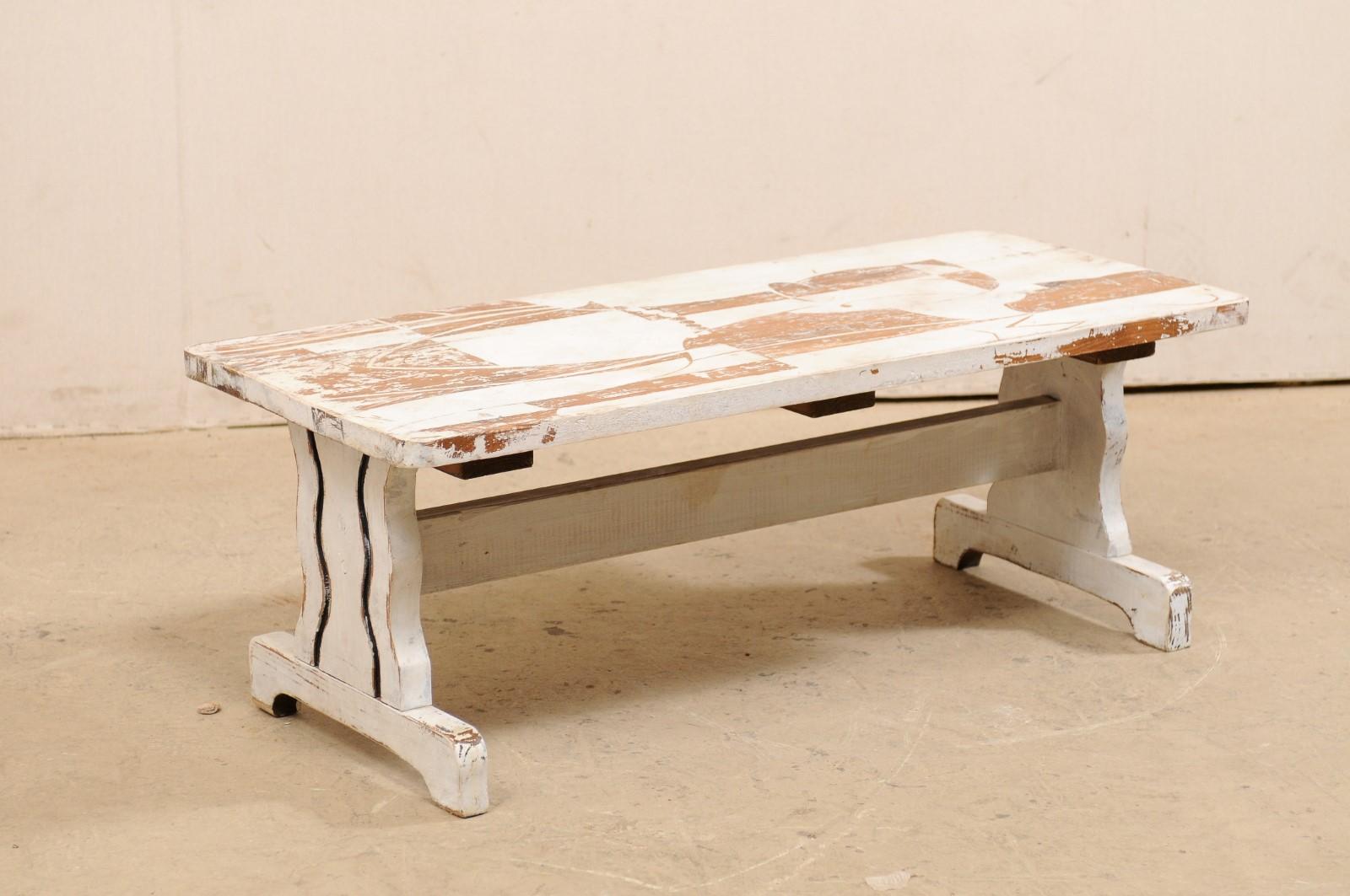 A European coffee table with an Art Deco themed painted top from the mid-20th century. A vintage coffee table from Europe has a rectangular-shaped top, just shy of four feet in length, which is supported by a pair of trestle legs (with curvy