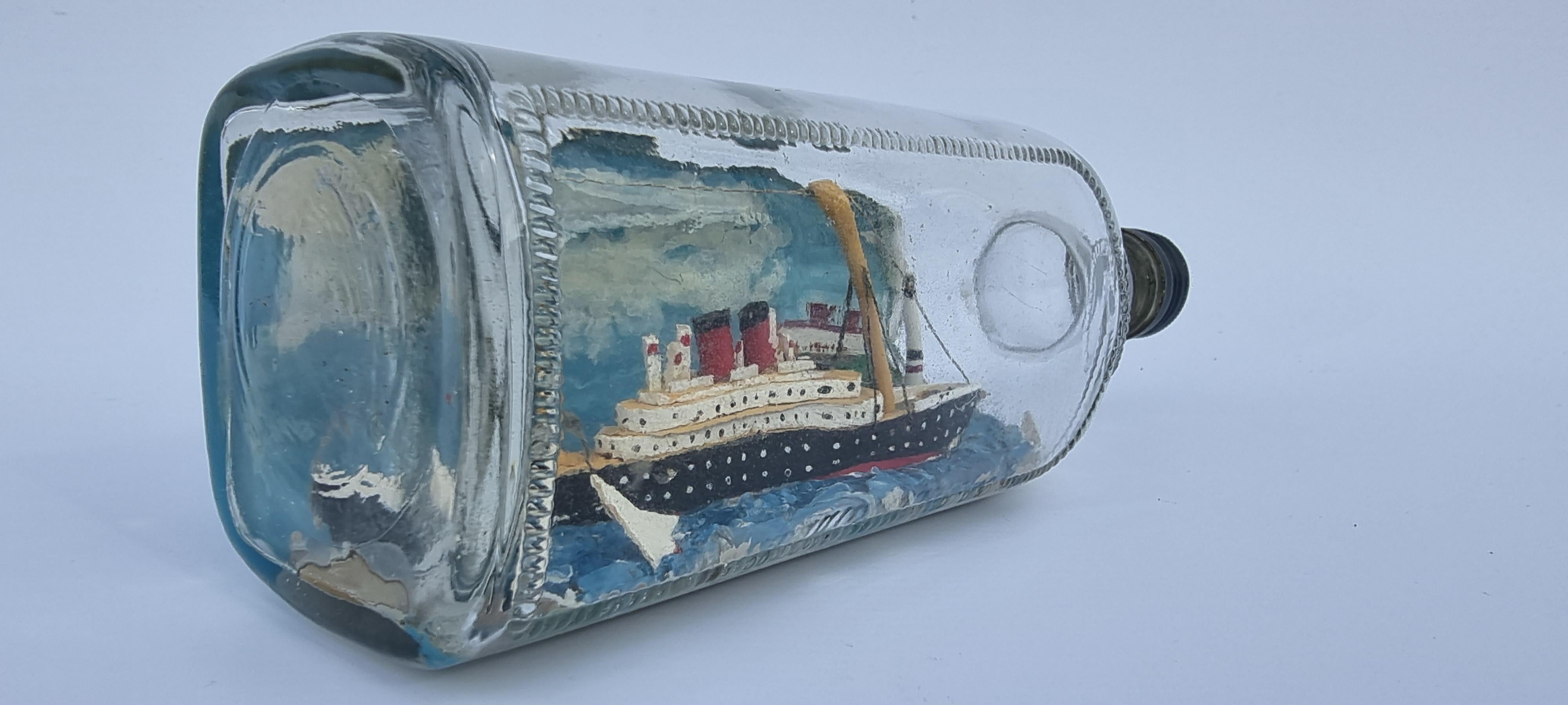 This early 20th century ship in a bottle diorama is of a rarer subject being an early large steam ocean going passenger liner set in a scene just off the shoreline with a lighthouse and cottages. The ship is on a choppy sea with two racing yachts in