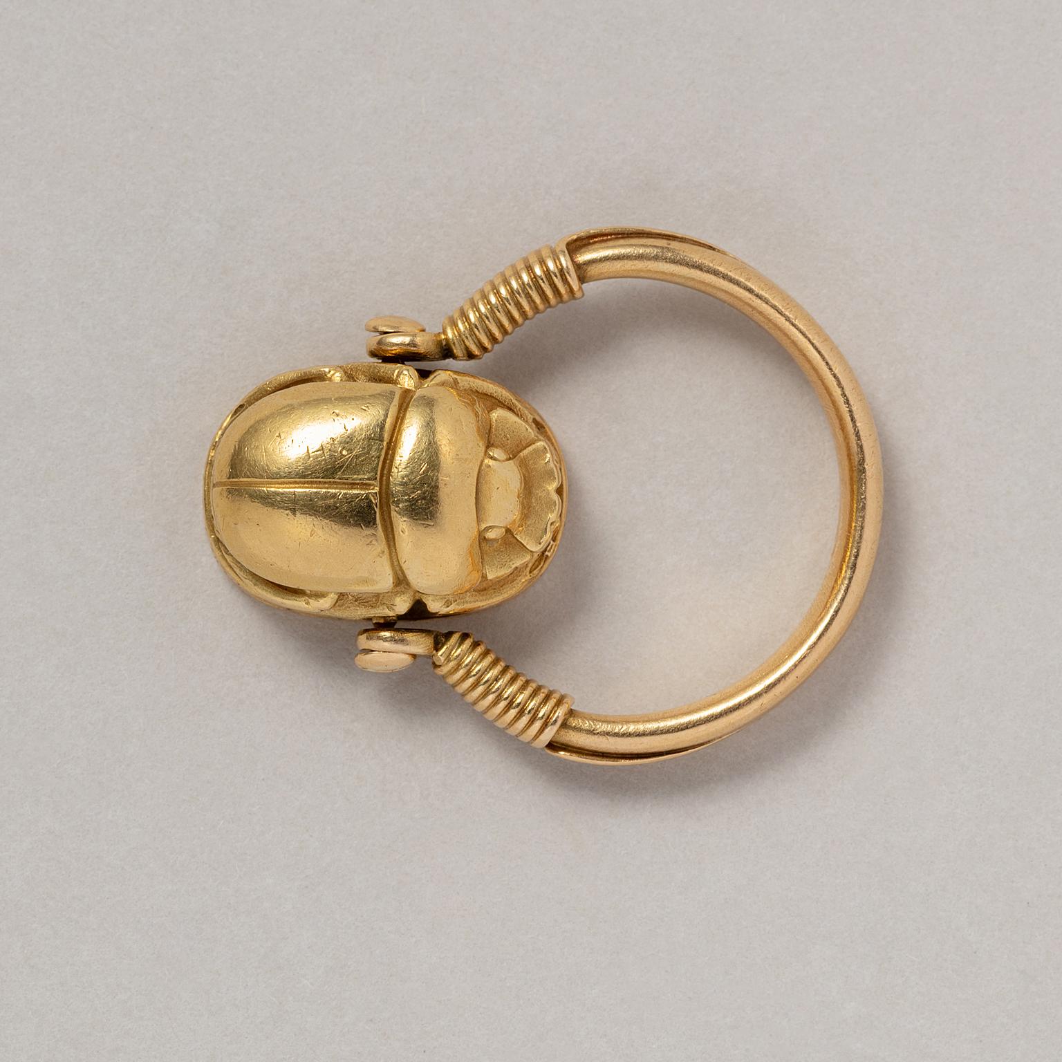 An 18 carat gold scarab ring swivel set with a rotating stylised Ancient Egyptian scarab between tapering shoulders wrapped in gold wire, with a rounded shank, the reverse of the scarab is blank, opening to reveal a hidden locket compartment, French