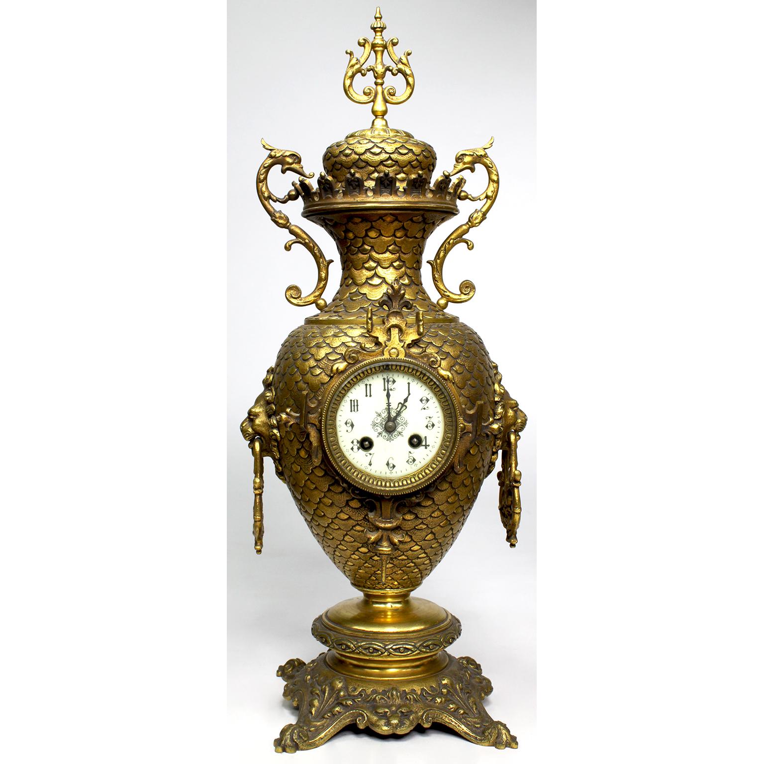 Horloge et chandelier en métal doré en trois parties, dans un style chippendale-arabesque. L'horloge en forme d'urne avec un corps festonné flanqué de poignées en forme de masque de lion et de décorations en volutes, l'horloge à double mouvement de