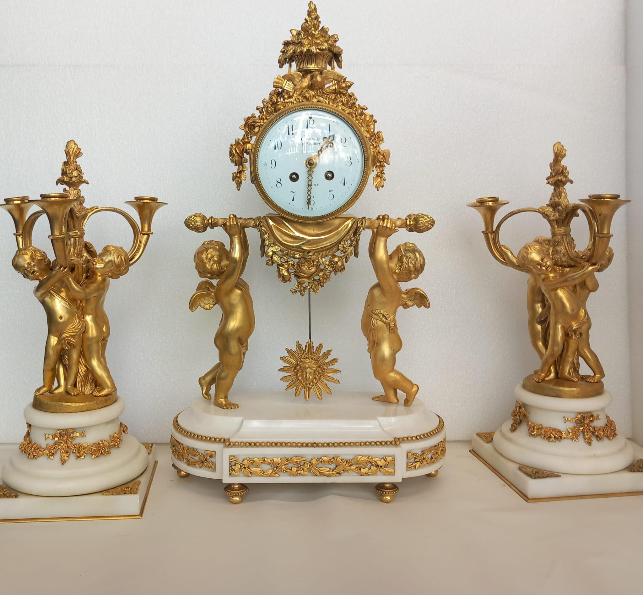 A French 19th Century gilt bronze clock garniture. The clock has two cherubs, holding the clock, on a white marble and Ormolu mounted base. The white enamel has Arabic numbers and chased gilt hands. The side pieces are Ormolu candelabras, cast in