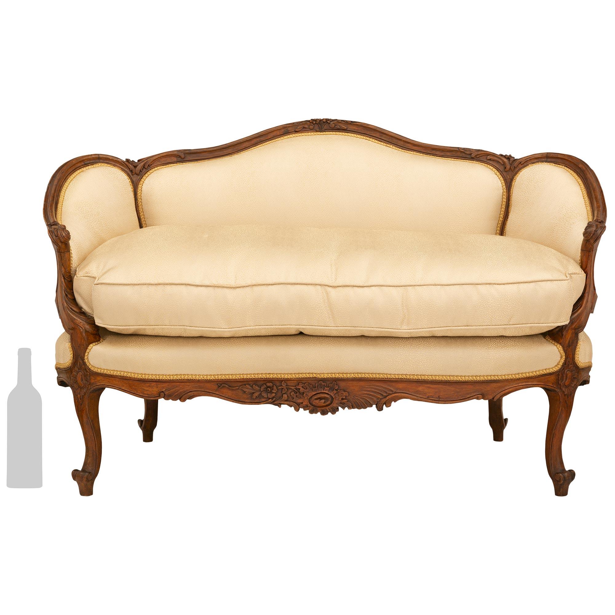 A charming and small scale French 19th century Louis XV st. Walnut settee. The settee is raised on elegant cabriole legs with a carved rosette below the scalloped frieze centered by a richly carved floral cabochon. Each upholstered scrolled arm is