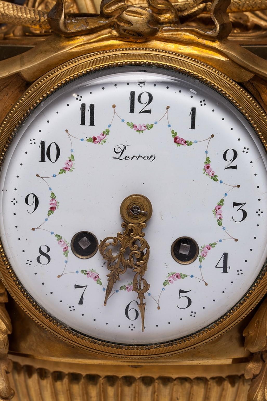 The mantel clock is composed of a circular, white enamel dial, featuring Arabic Numerals, set within a vase-form case. The clock is on an oval base which is on four legs. The mechanism is located on top of a column souraunded a laurel garland. A