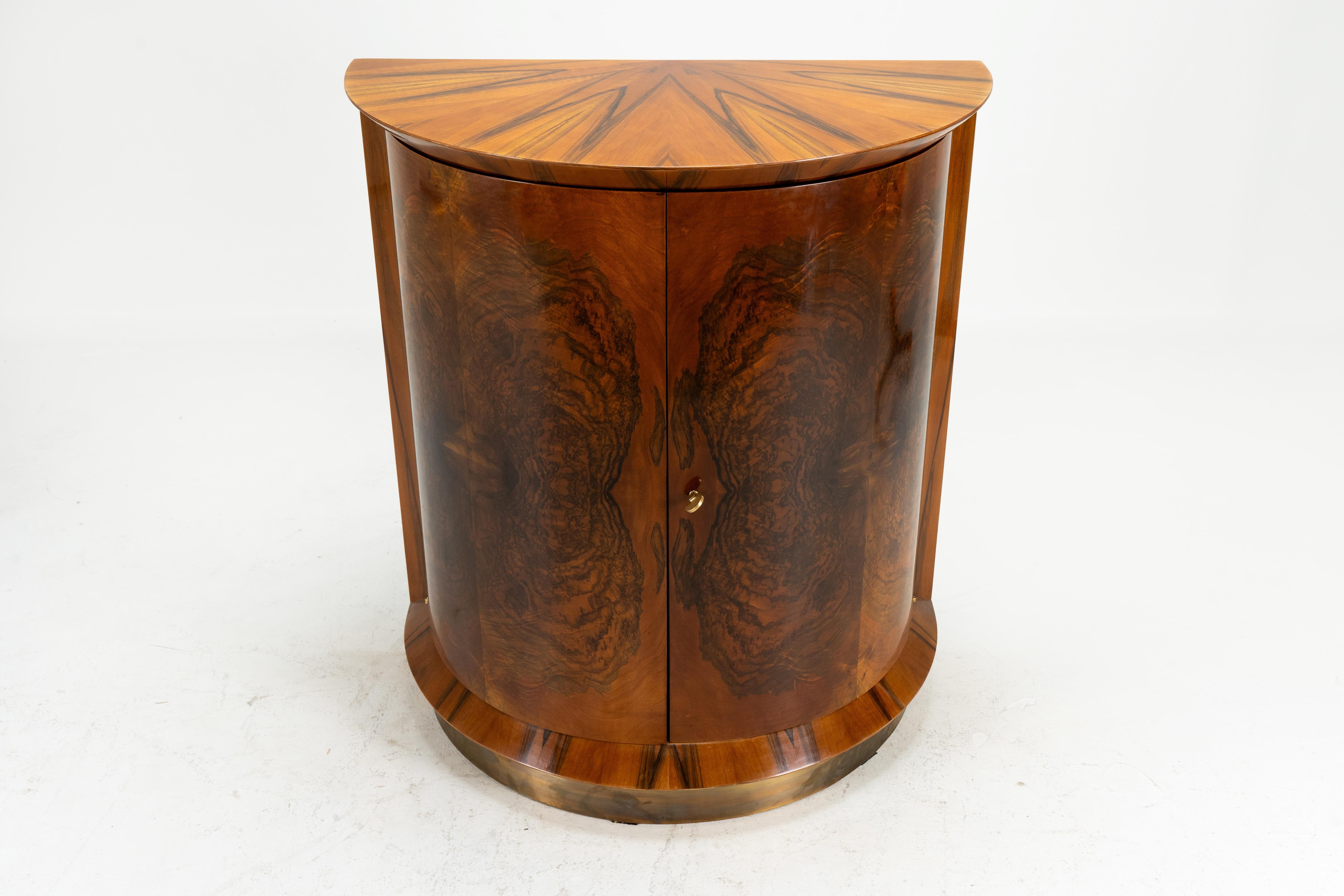 This elegant semi-circular bar cabinet, which is clad on the outside with book-matched walnut burl veneer, rests on a base clad in patinated brass. The finish is silky and smooth French polish.  The top veneers are of a pie-wedge design radiating