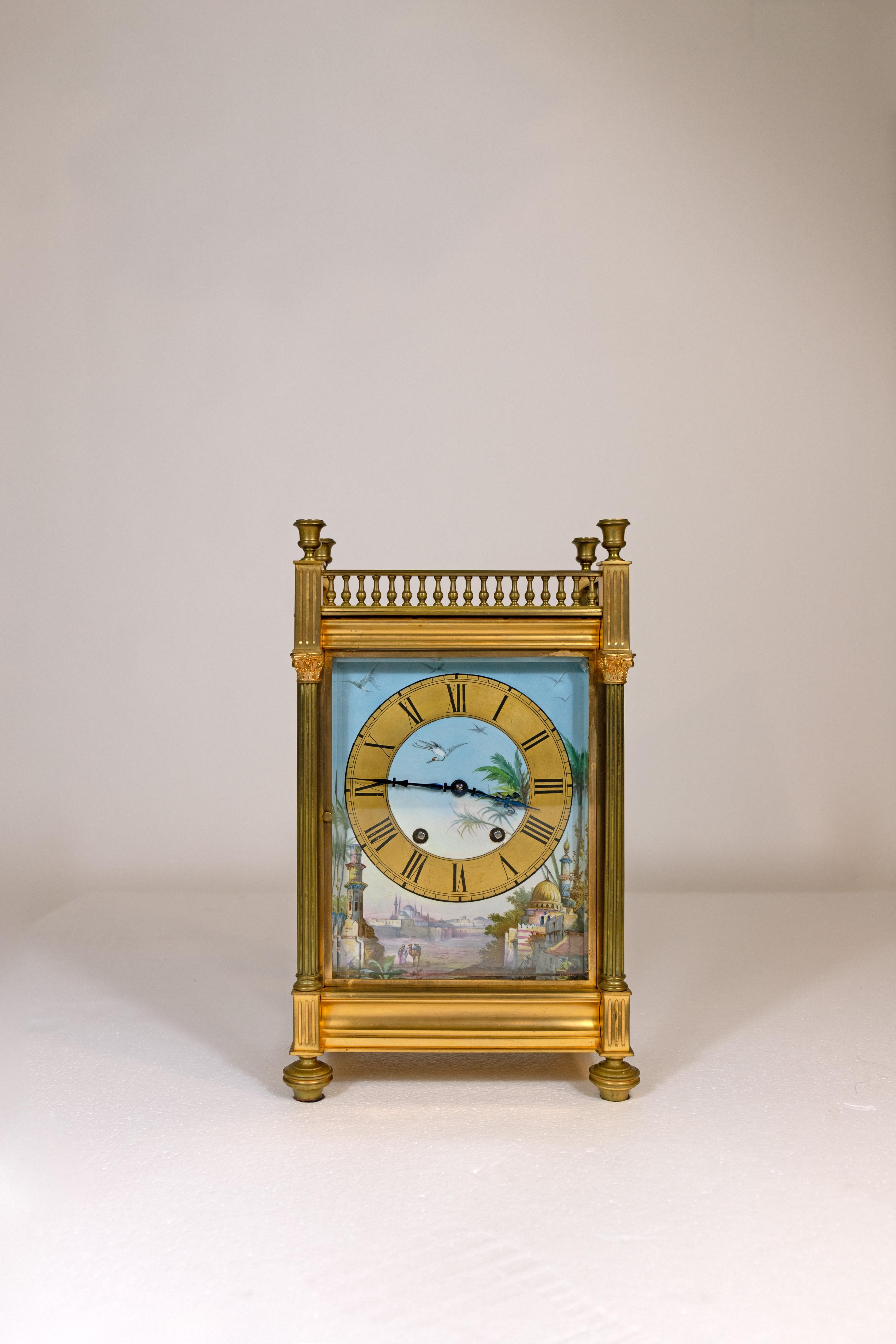 A mantel clock in gilt-bronze and glass featuring an architectural case with neoclassical detailing. The four corner columns are topped with urn finials and joined by a pierced balustrade gallery. Each side is framed in beveled glass, offering a