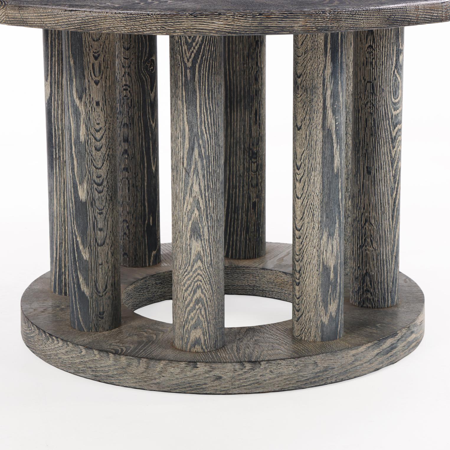 Escultural mesa de comedor redonda francesa de roble cerusado, con tablero circular elevado sobre ocho soportes cilíndricos unidos por una base circular plana. El acabado cerámico, cálido y texturizado, resalta el veteado natural del roble,