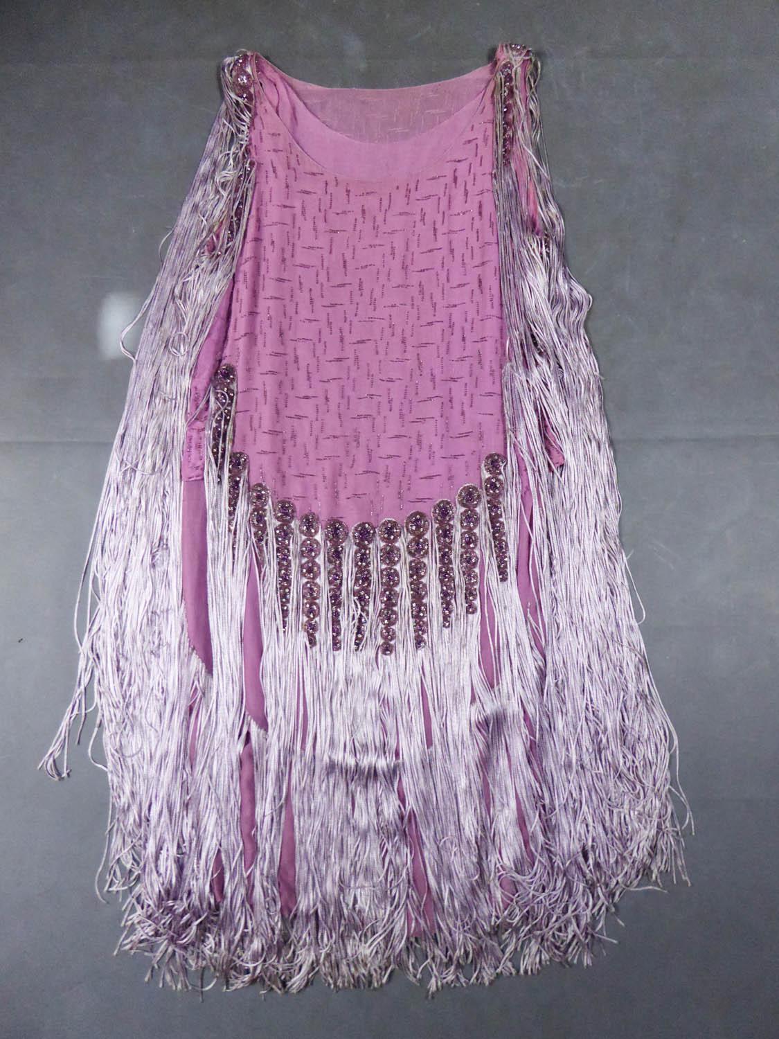 Circa 1928
France

Beautiful Charleston-type ball gown dating from the Twenties . Tubular dress in pink-purple silk crepe embroidered with geometric patterns in glass beads. Long skittles in silver cream silk laces starting from the shoulders and