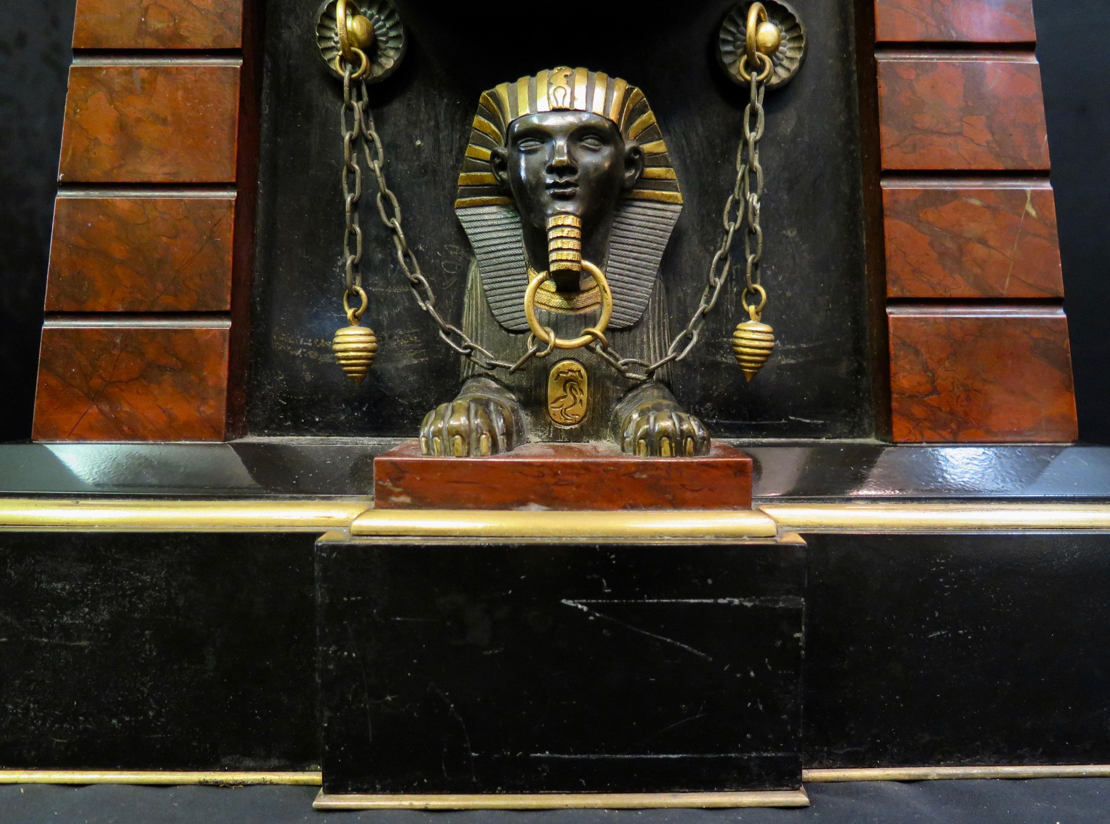 L'horloge a un boîtier trapézoïdal bombé, un dôme avec un relief en bronze sur le devant représentant une déesse égyptienne Isis partiellement dorée, flanquée de cobras et de volutes de lotus, des colonnes nervurées en marbre rouge sur les côtés, un