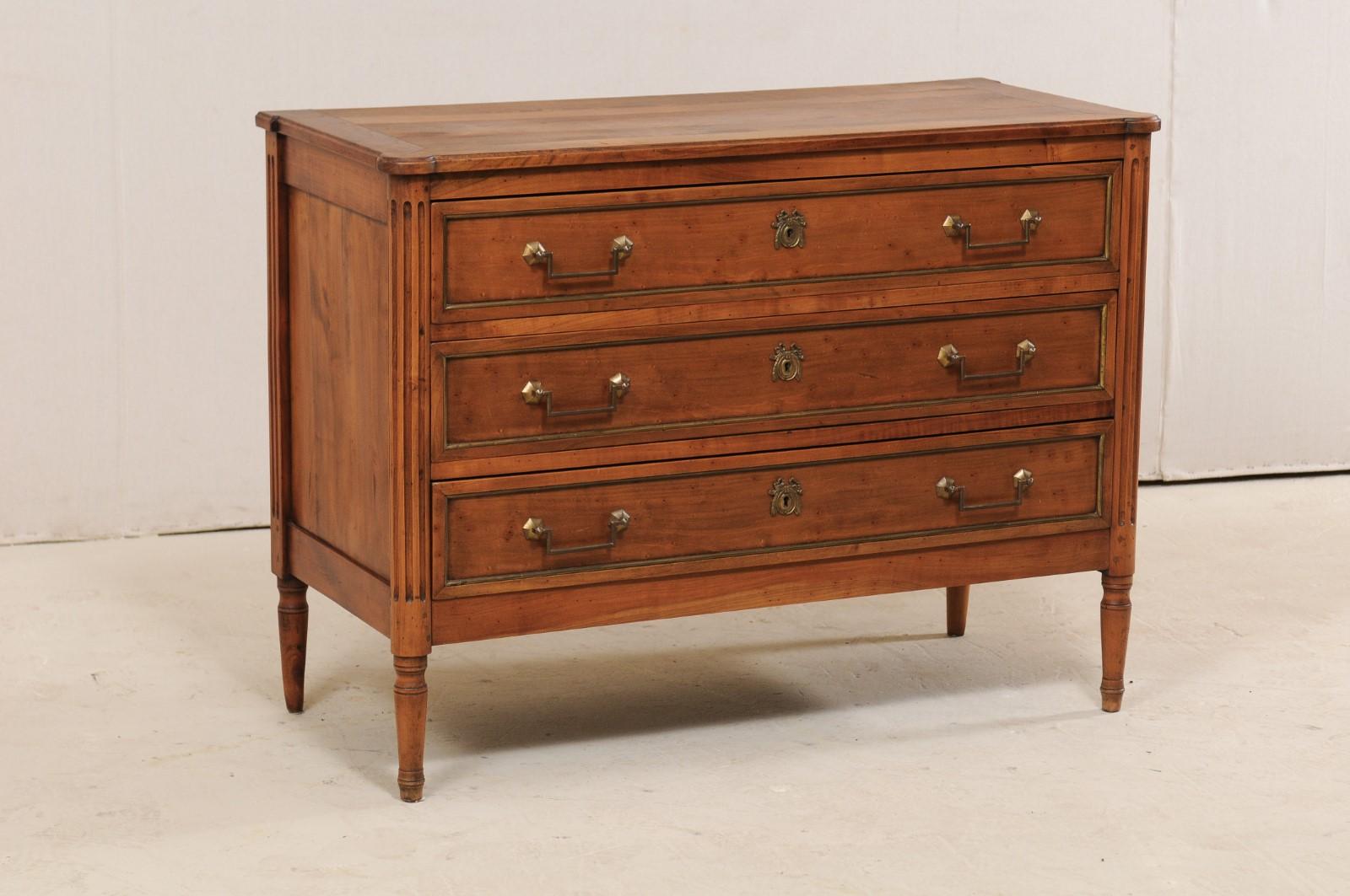 A French chest of three drawers with fluted details and brass trim from the early to mid 20th century. This beautiful fruit-wood commode from France, designed in Louis XVI style, features a slightly overhanging top with rounded, pronounced