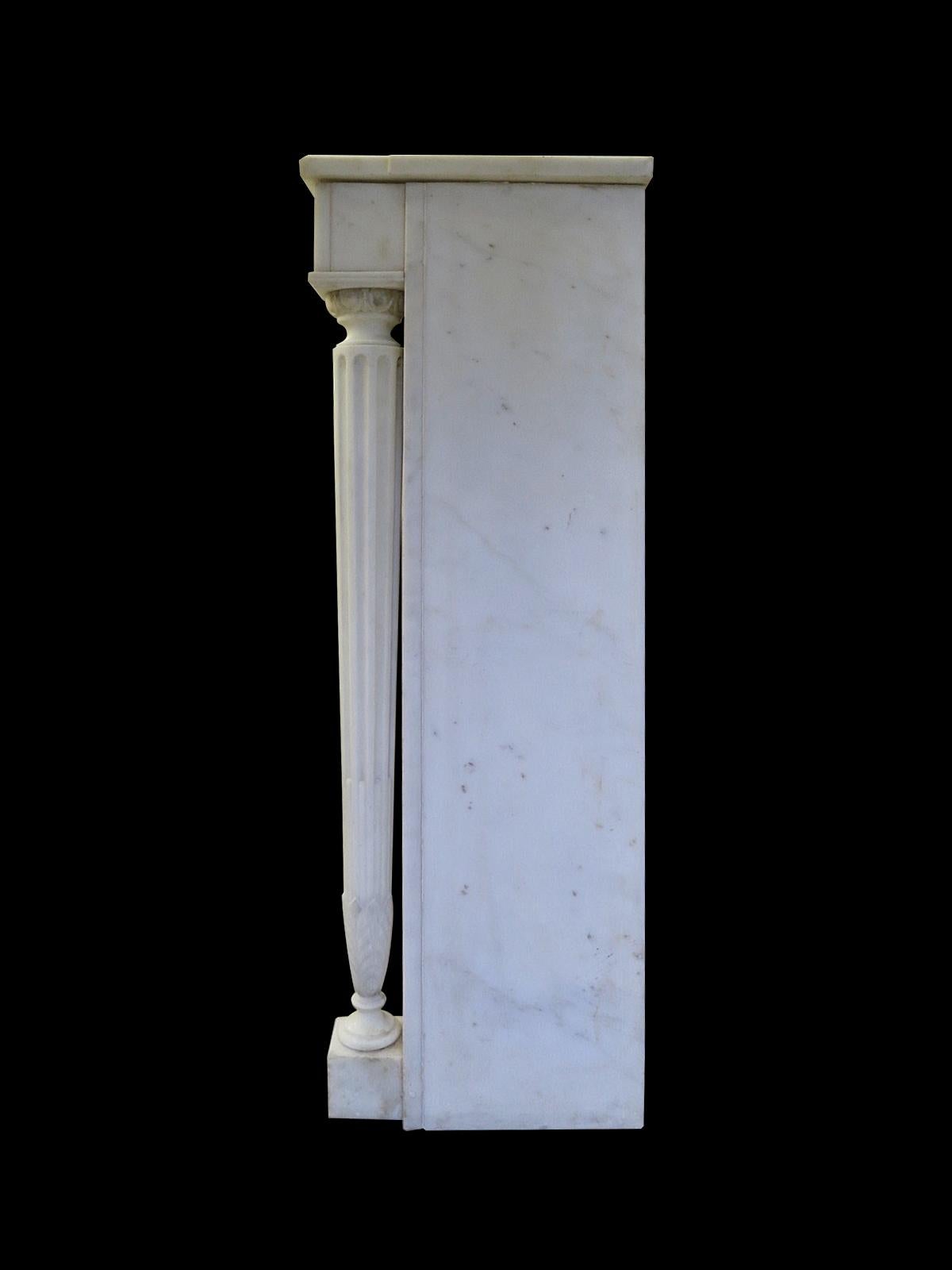 A late 19th century Louis XVI style fireplace in white marble, with delicately tapering and fluted fully disengaged columns terminating in Acanthus leaf, and capped with egg and dart. The columns supporting a fielded paneled frieze, with carved
