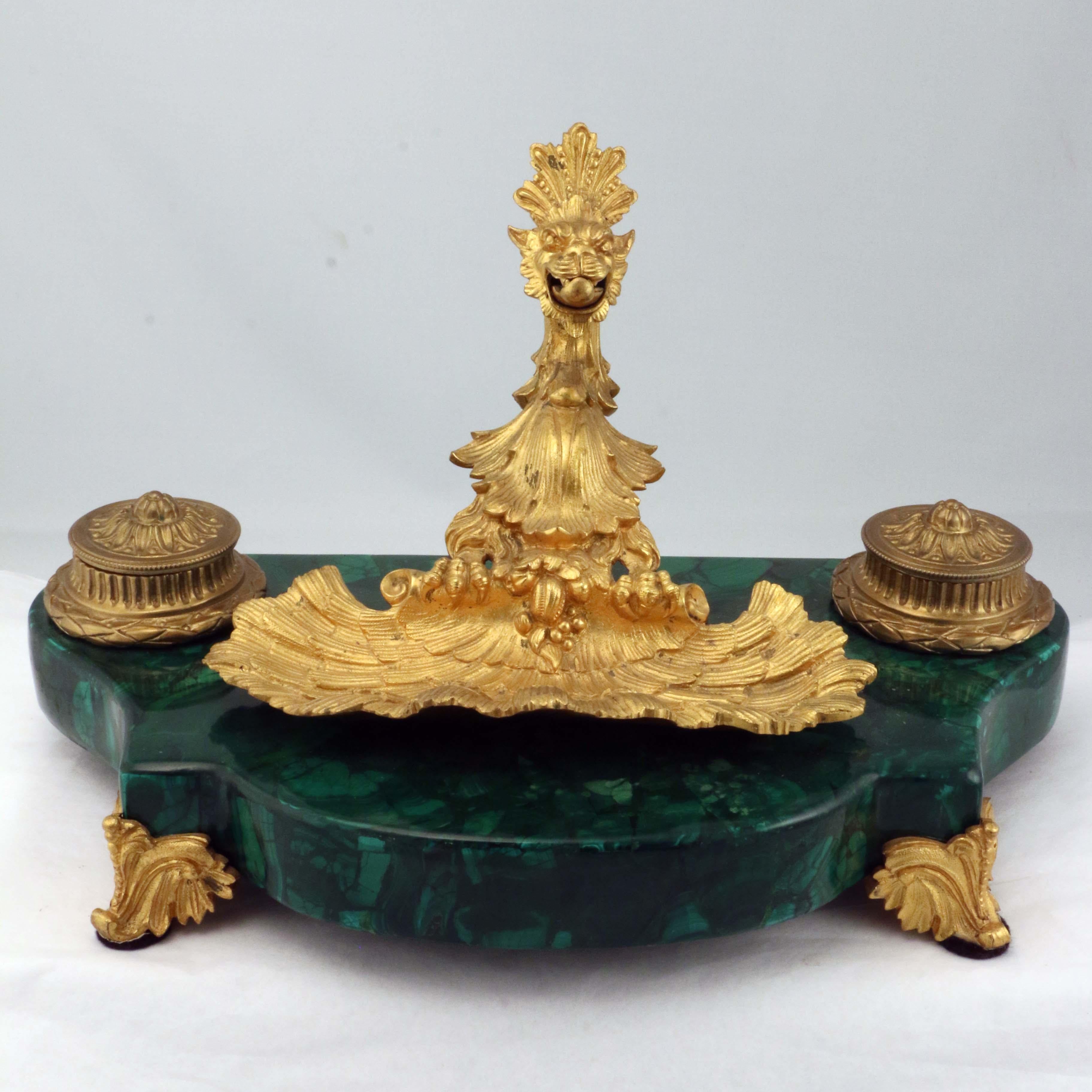This luxurious encrier de bureau is raised on scrolling and leafy feet mounting a malachite-finished base. The tray itself is modelled as a shell rising to a spring-loaded pen holder. The animal grips the pen between its teeth. The flanking covered