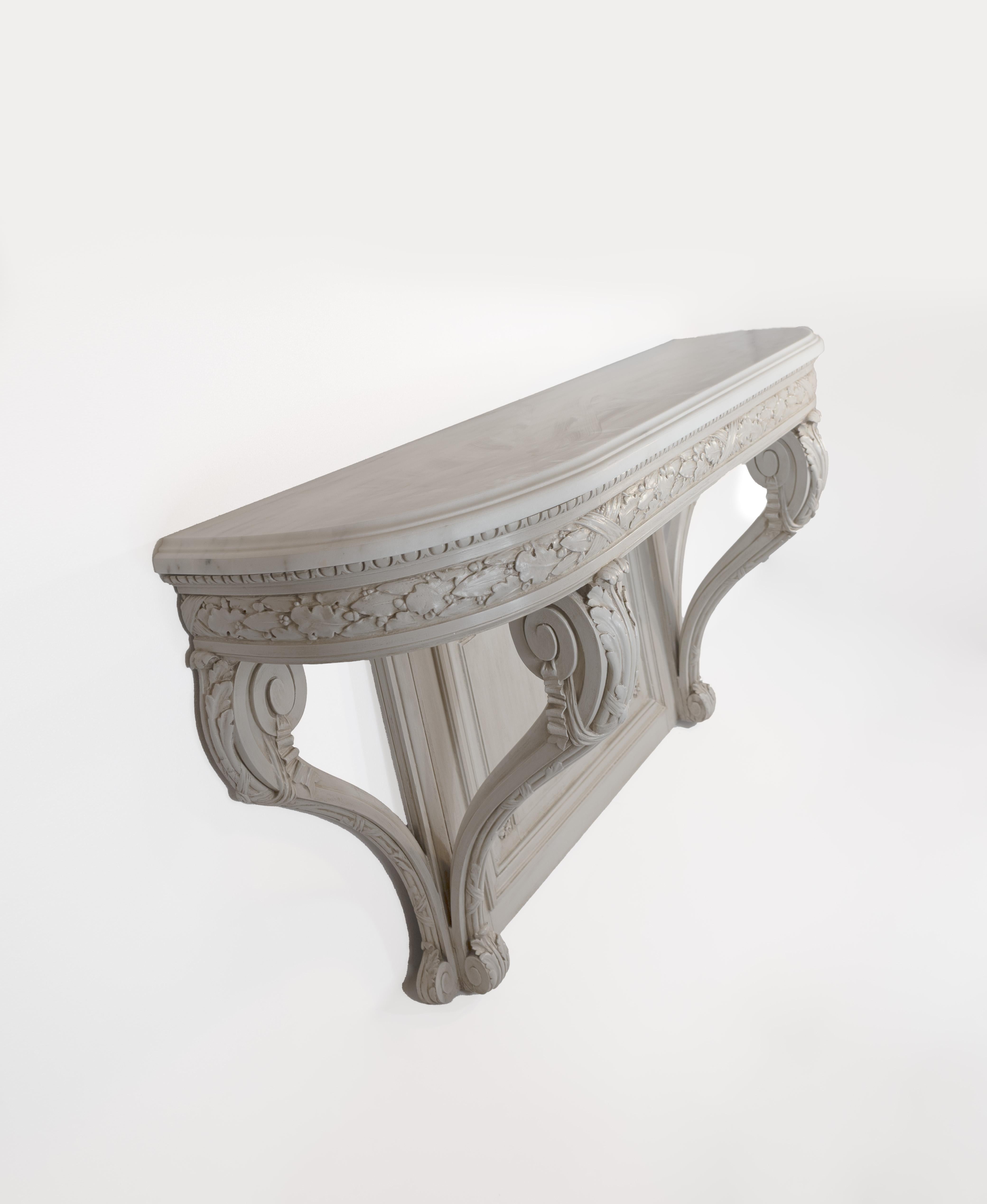 This sculptural wall-mounted console table is made from carved wood and finished in a layered ivory-toned patina; it features a gracefully shaped marble top over a richly detailed frieze carved with oak leaves and acorns in crossed ribbon bands. The