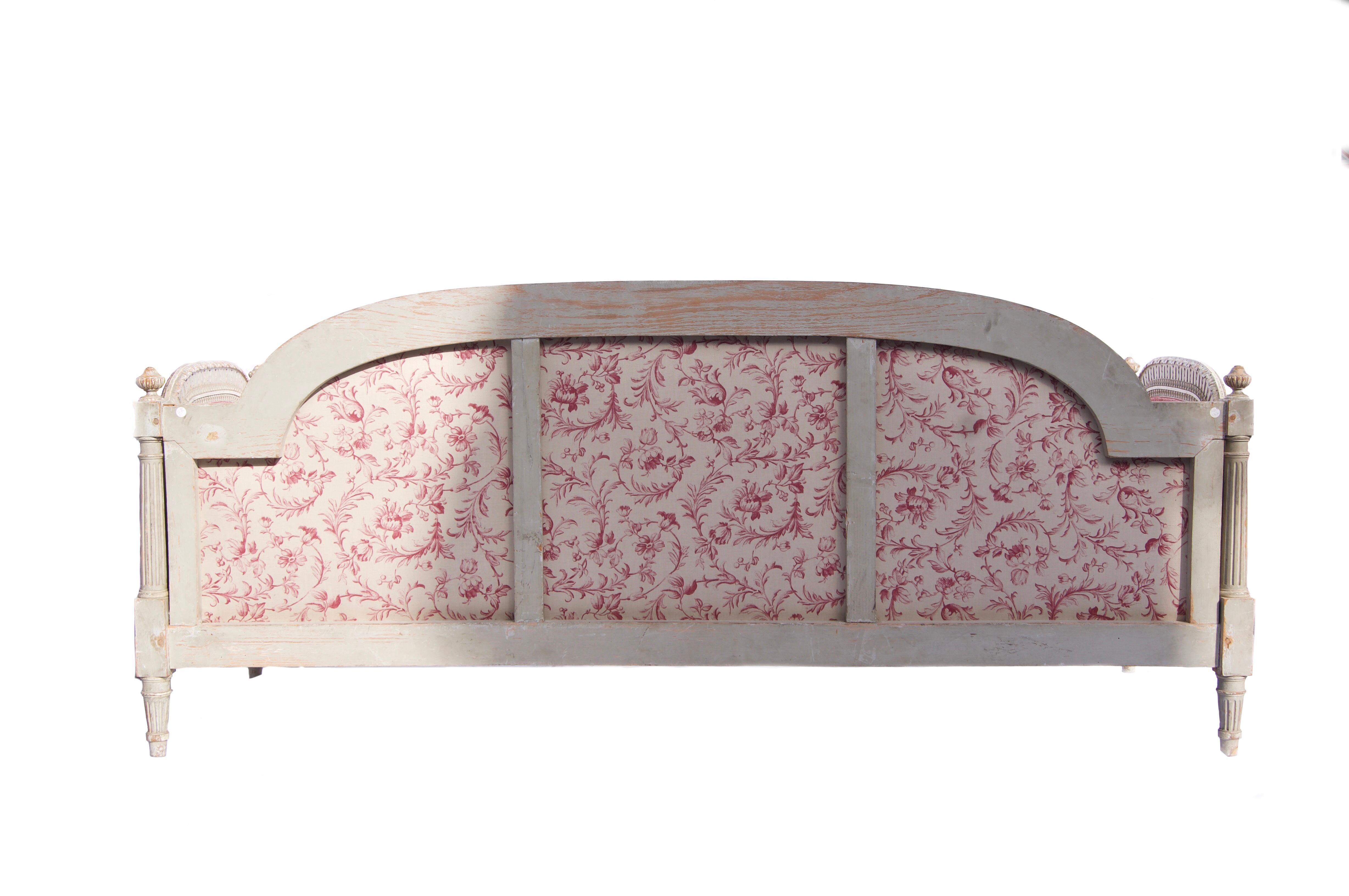 A French Louis XVI style painted and carved wood daybed, late 19th century with shaped back and sides with knop finials, grey paint, upholstered in a raspberry fabric, with a pair of bolster cushions.