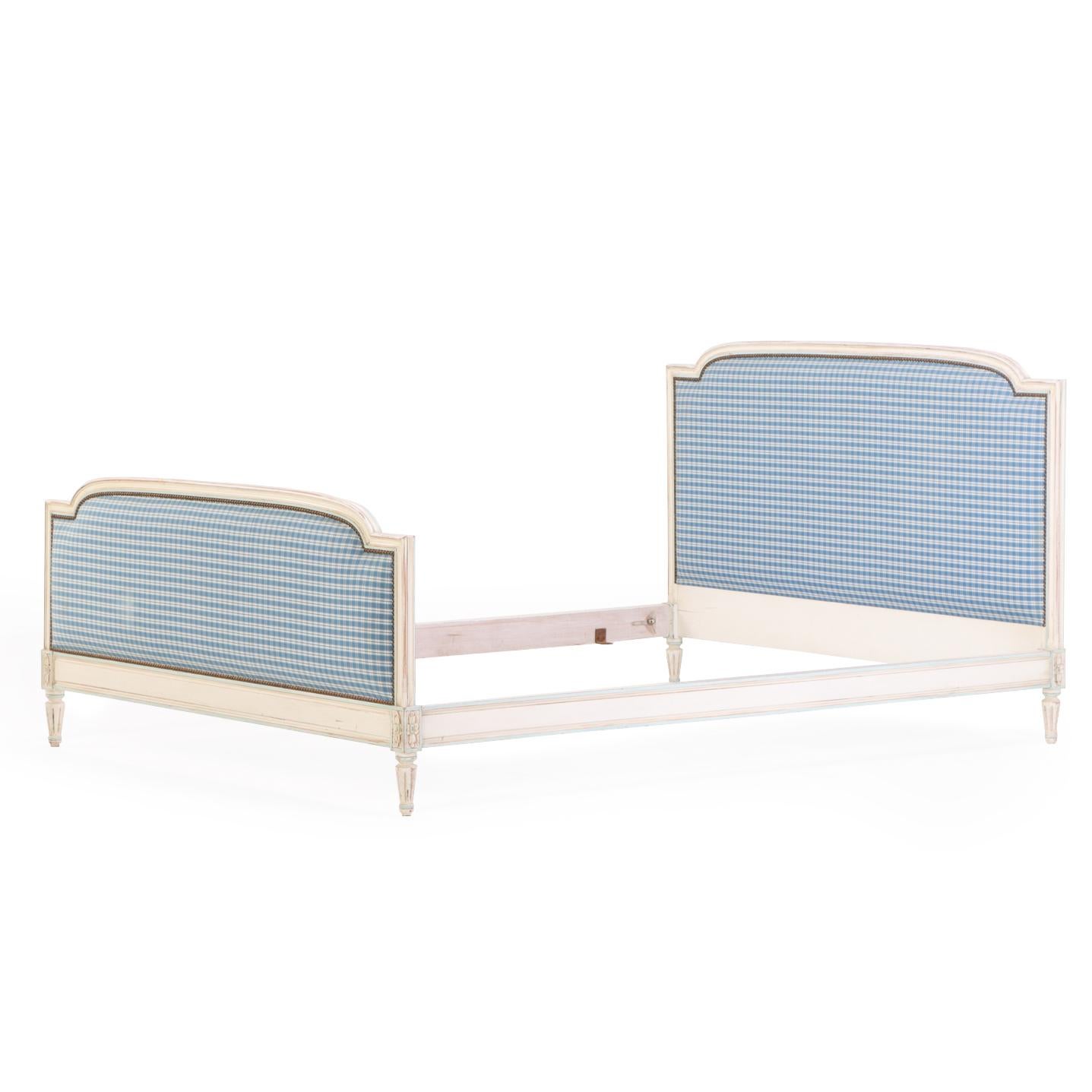 A charming and finely detailed bed featuring carved rosette details, turned tapering feet, and upholstered headboard and footboard in a classic blue and white checked fabric. The frame is finished in a soft, vintage white paint, lightly worn to